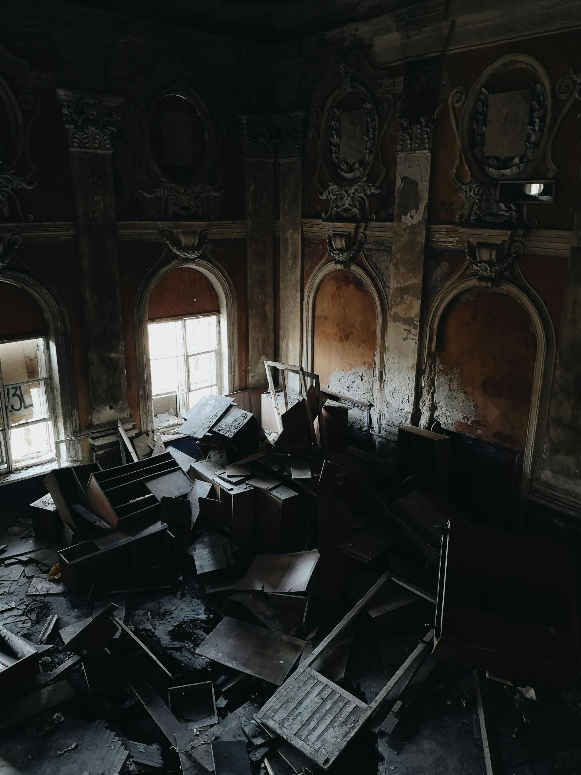 Interior of a dilapidated building, strewn with debris. Dark, crumbling walls surround windows, and fragments of furniture litter the floor.