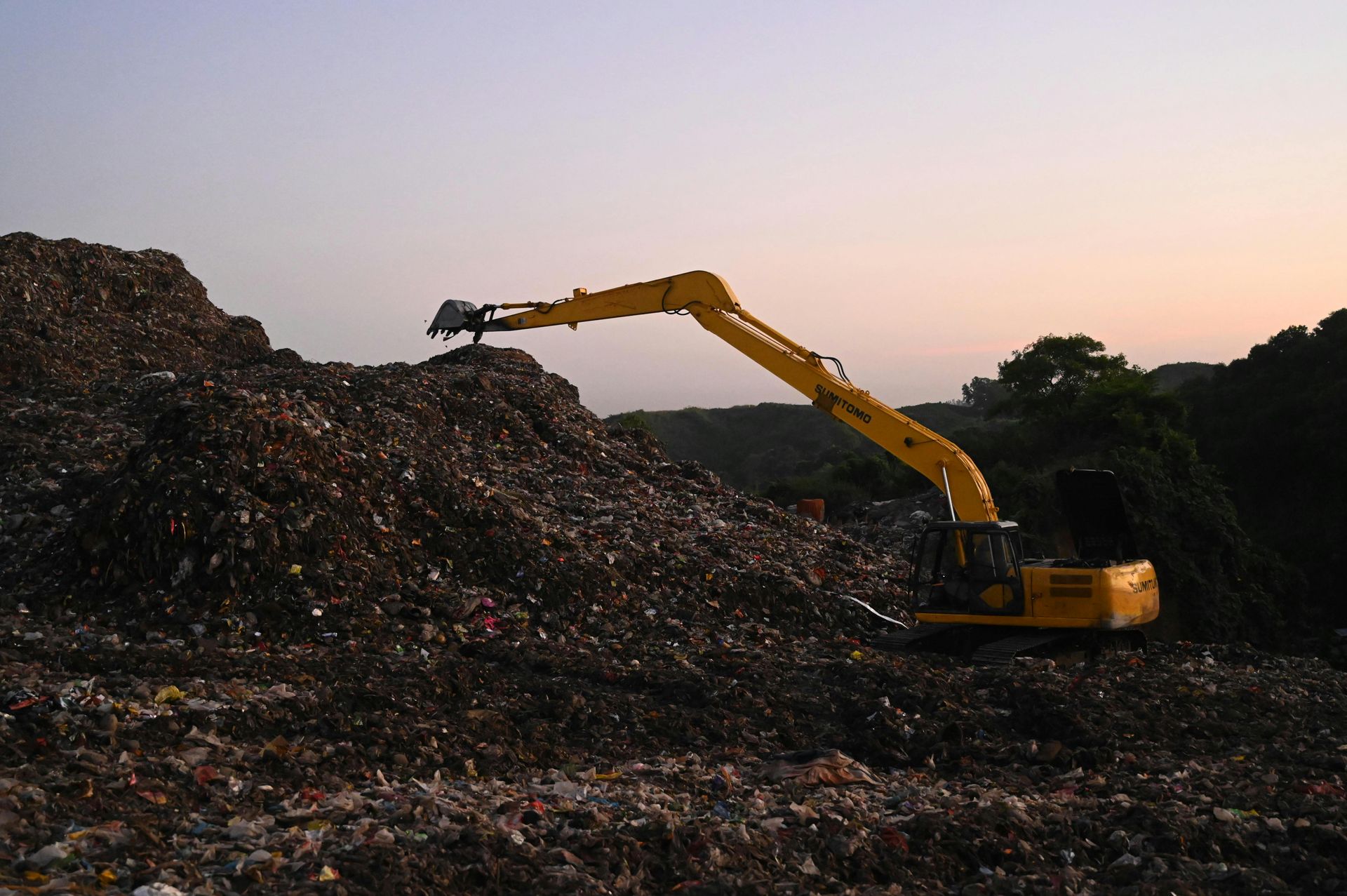 Yellow excavator working on a large pile of waste, likely in a landfill, at dusk.