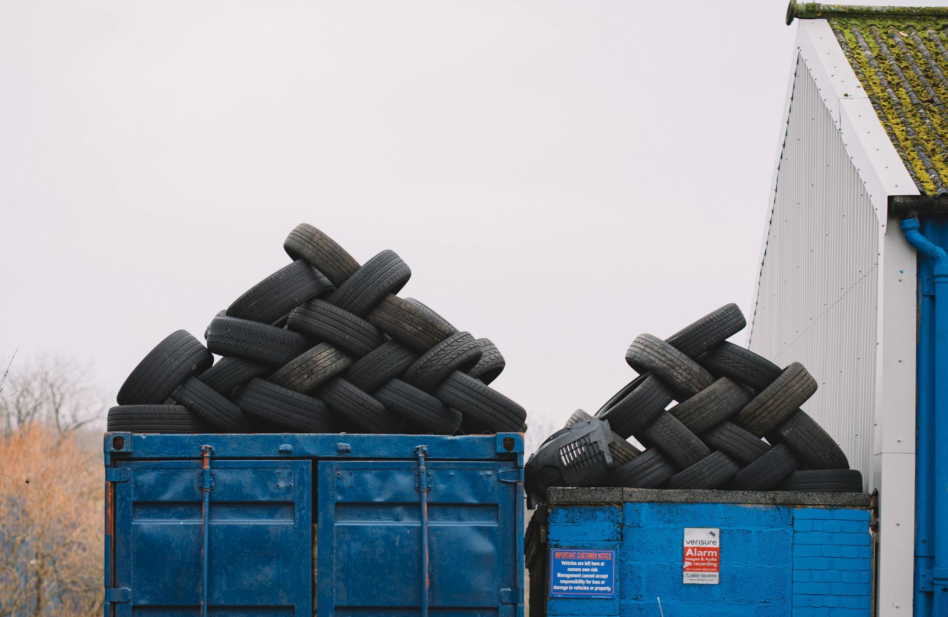 Two blue dumpsters filled with used tires, stacked in a weaving pattern. A white building and cloudy sky are in the background.