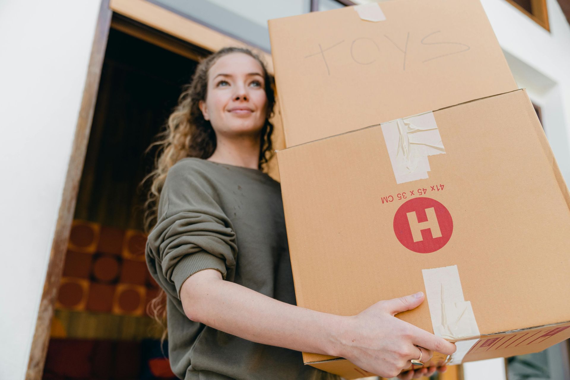 Woman with curly hair carrying two large cardboard boxes, possibly moving, smiling outside a building.