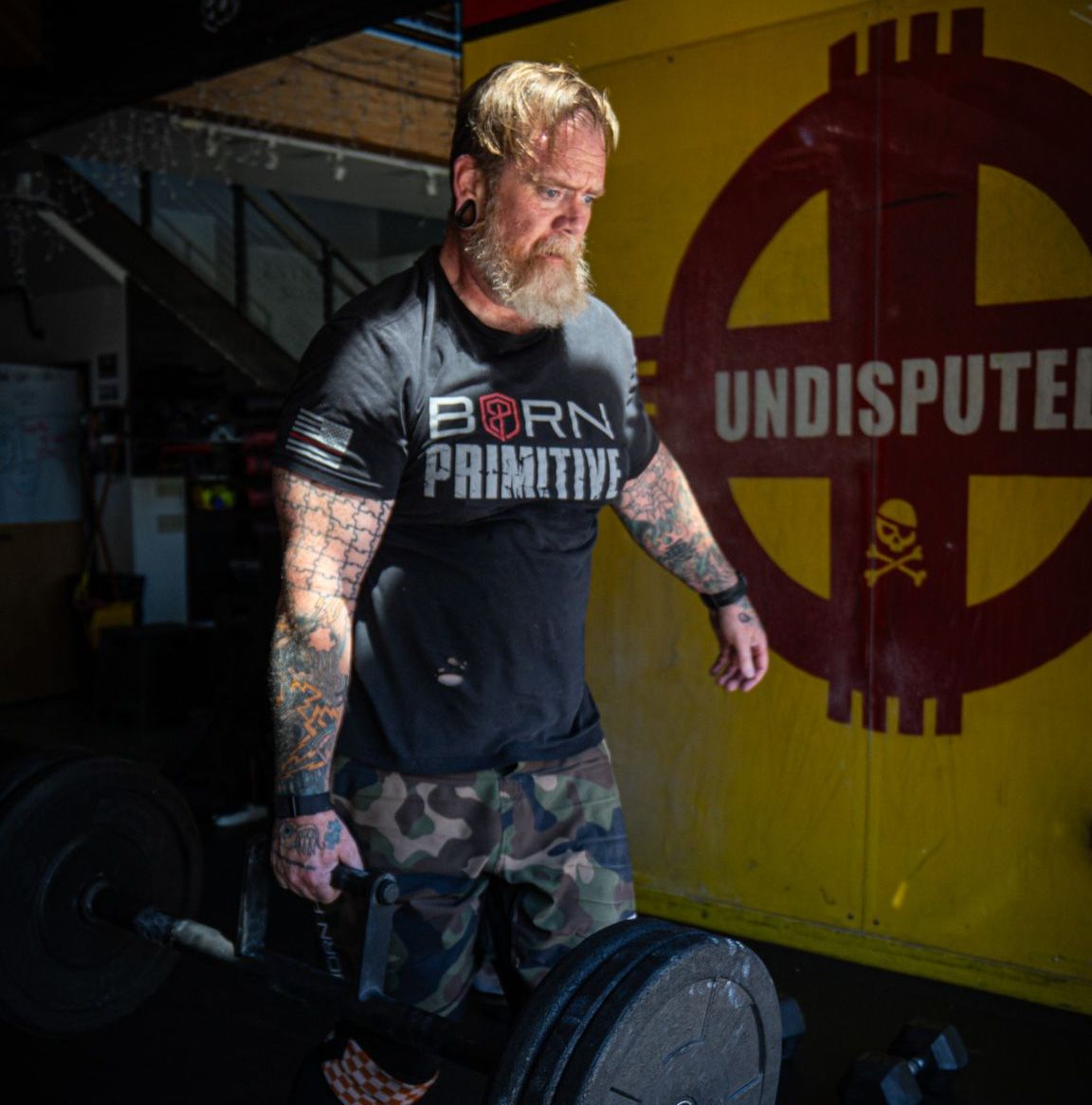 A man wearing a born primitive shirt is lifting a barbell