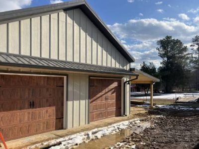 Garage with brown doors, beige siding, and dark gray roof, set in a snowy, muddy landscape.
