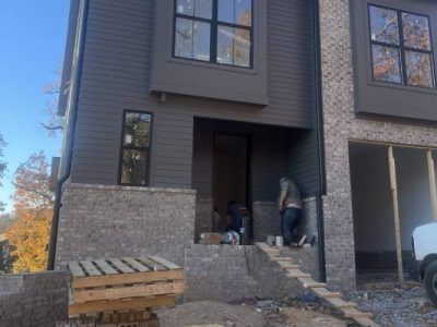 Construction workers installing brick on a two-story building's facade. Gray siding and windows, blue sky.