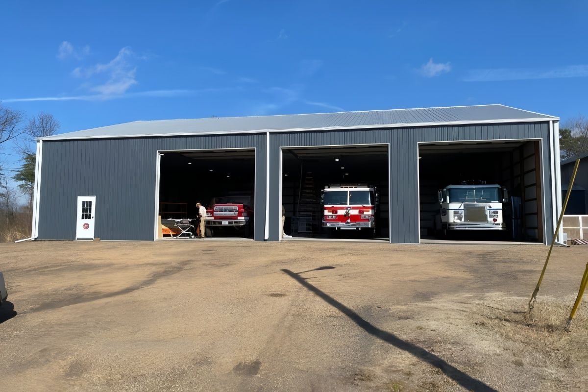 Three-bay fire station with fire trucks inside. Gray building with white door, under a blue sky.