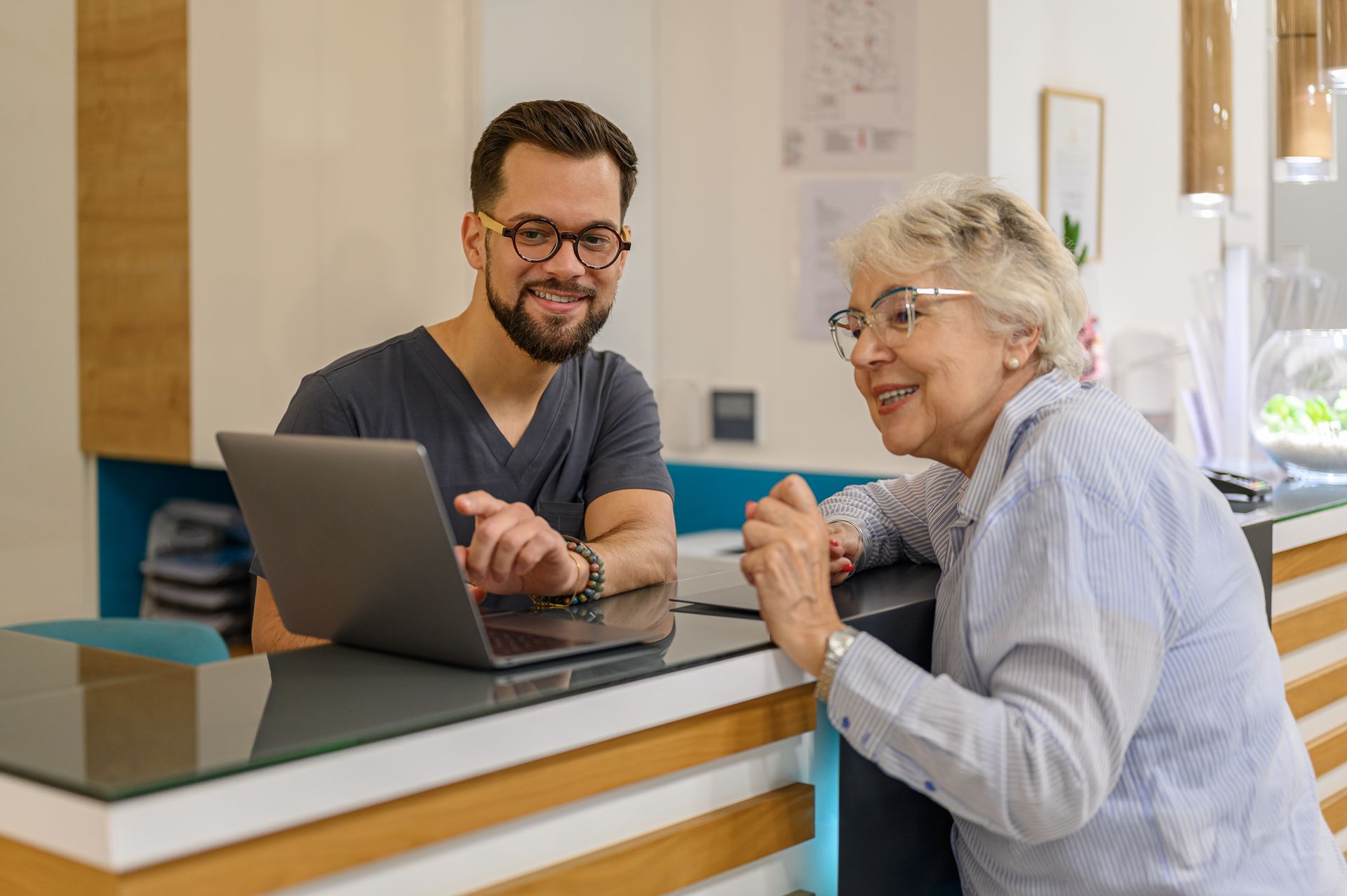 An eye doctor and senior woman collaborate at a clinic desk, focused on a laptop in front of them.