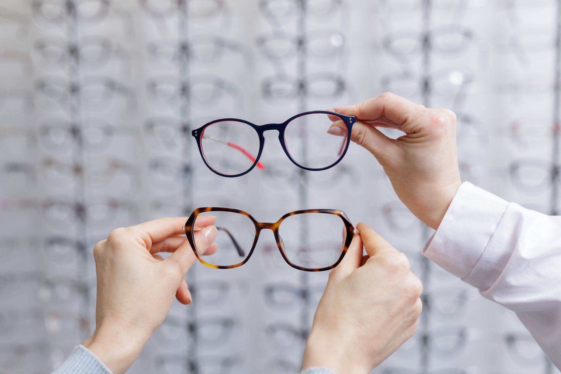Hands holding two eyeglass frames in front of an eye chart