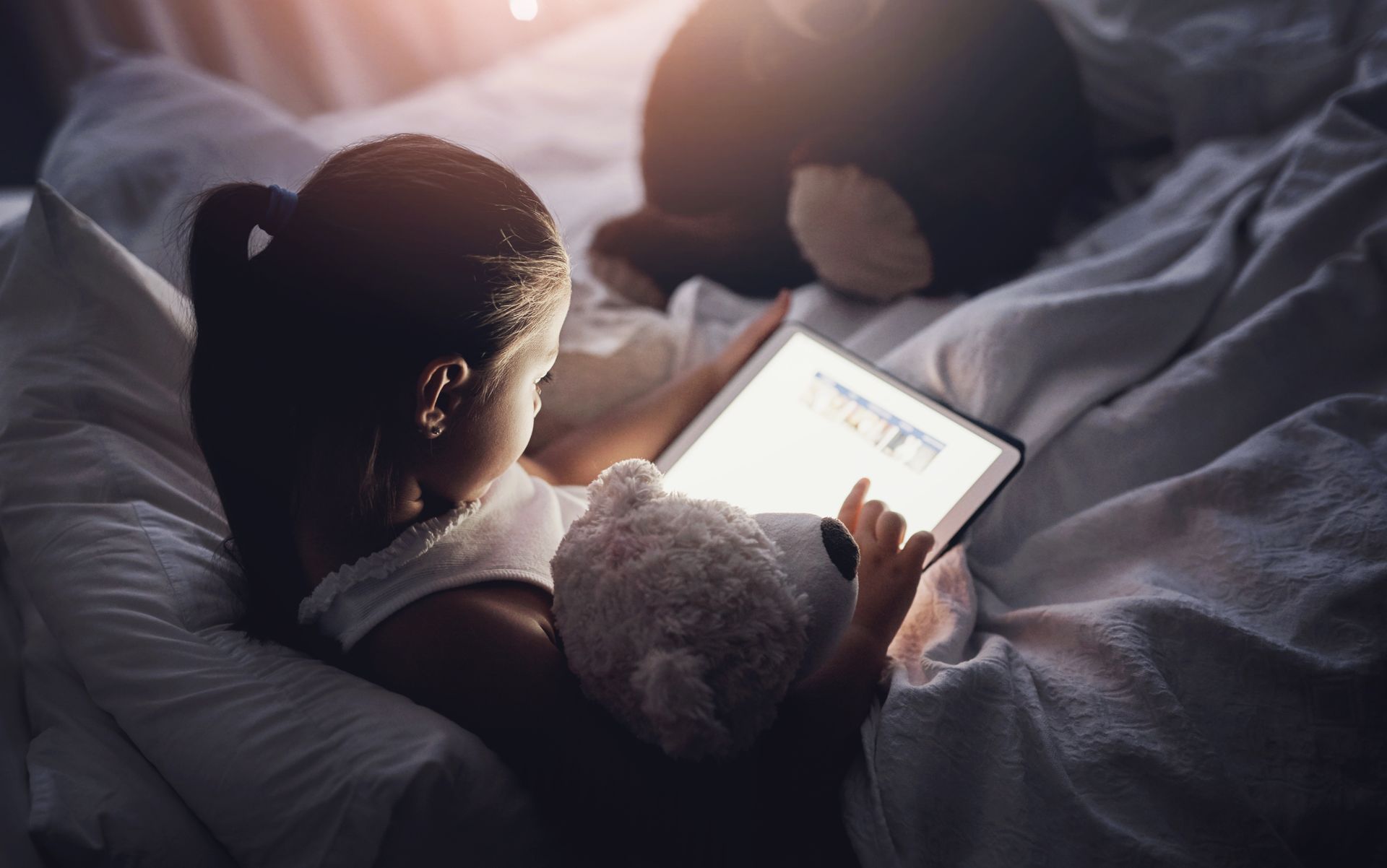 Person sitting in bed using a tablet beside a teddy bear in soft morning light