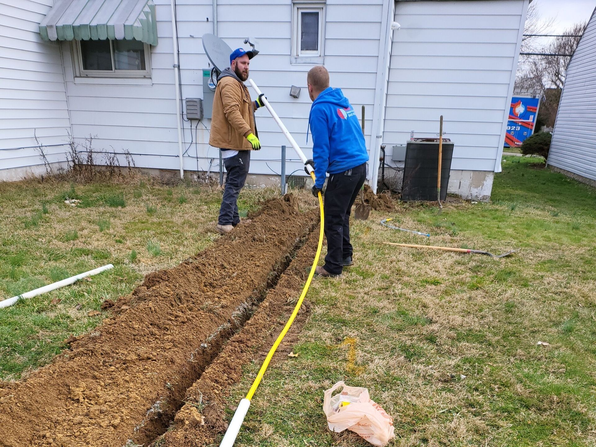 Two men are digging a hole in the ground in front of a house.