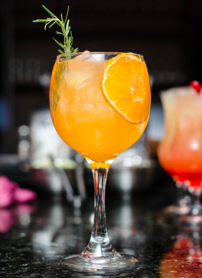 Orange cocktail in a stemmed glass with a rosemary sprig and citrus garnish on a bar counter