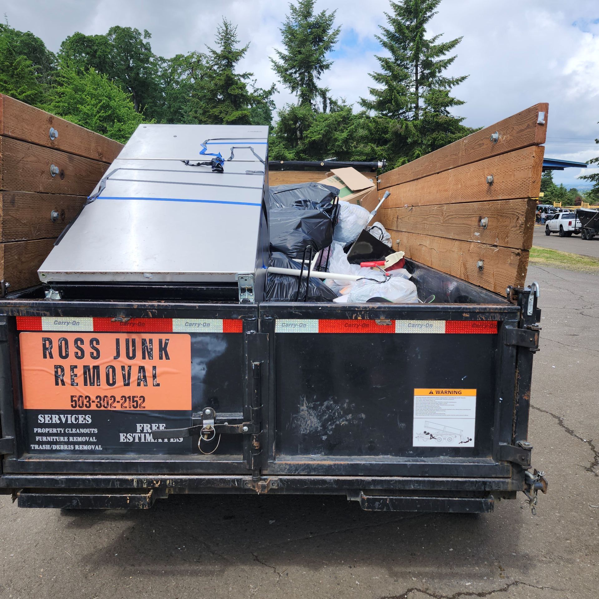 A black dumpster filled with junk, including an appliance. The dumpster has a Ross Junk Removal sign on it.