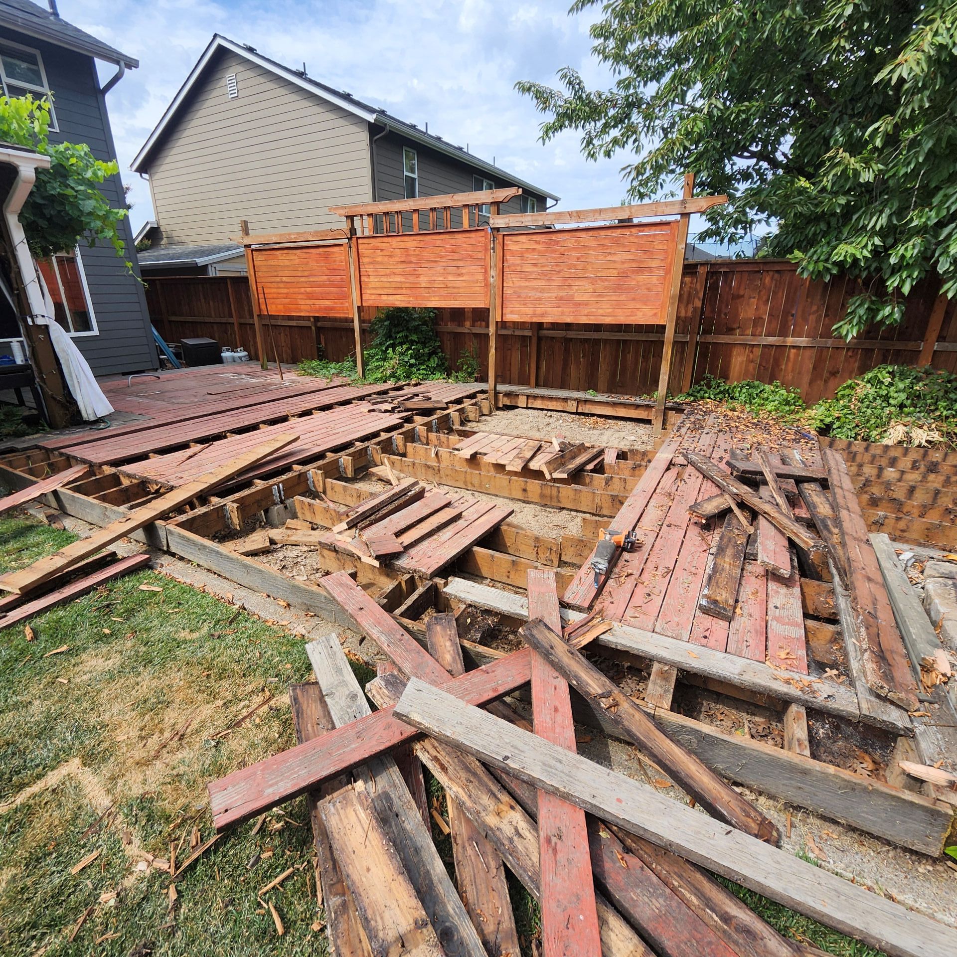 Deck demolition in a backyard; scattered wood planks on grass. Reddish-brown fencing and wooden privacy screen in the background.