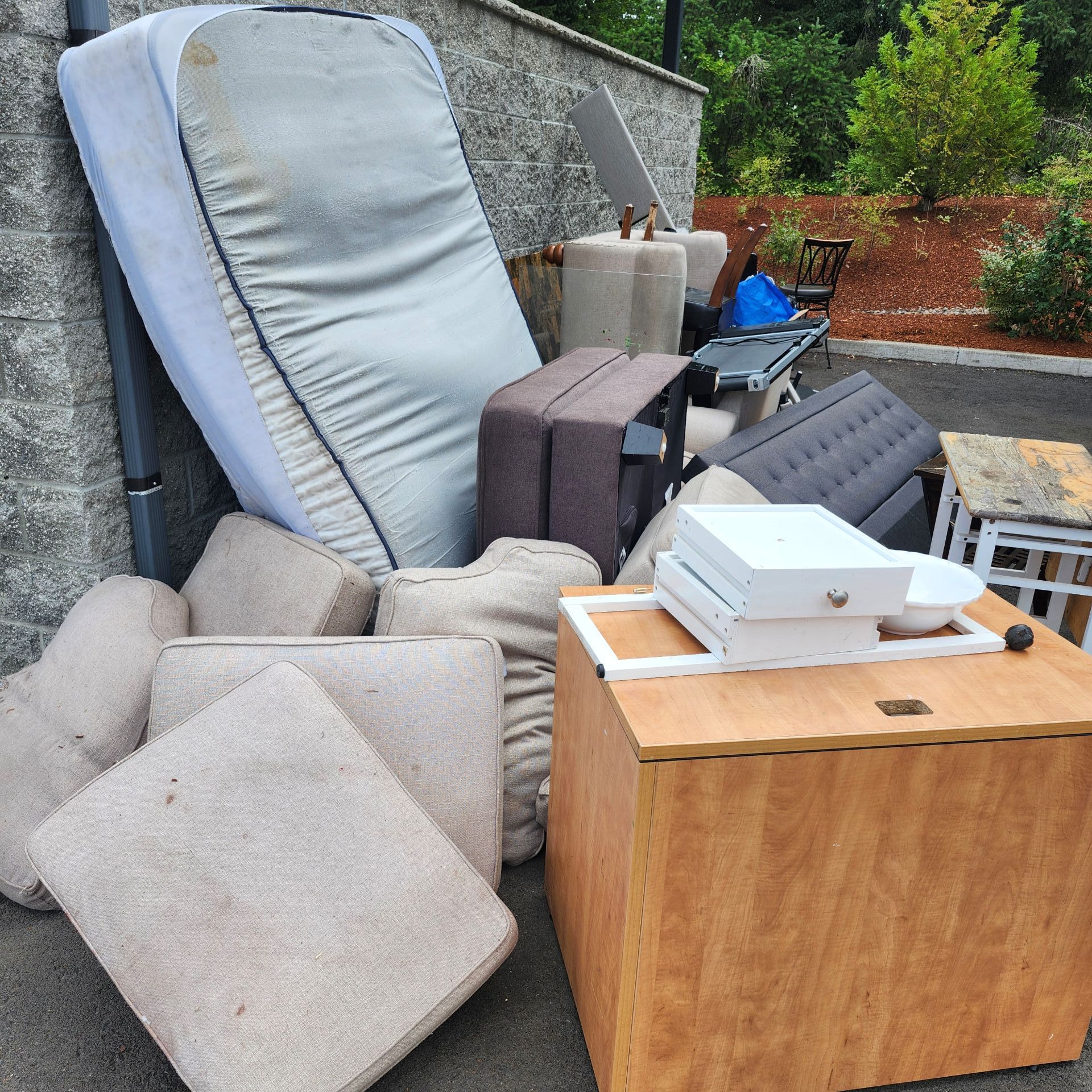 Pile of discarded furniture, including a mattress, cushions, and wooden cabinet, outdoors.