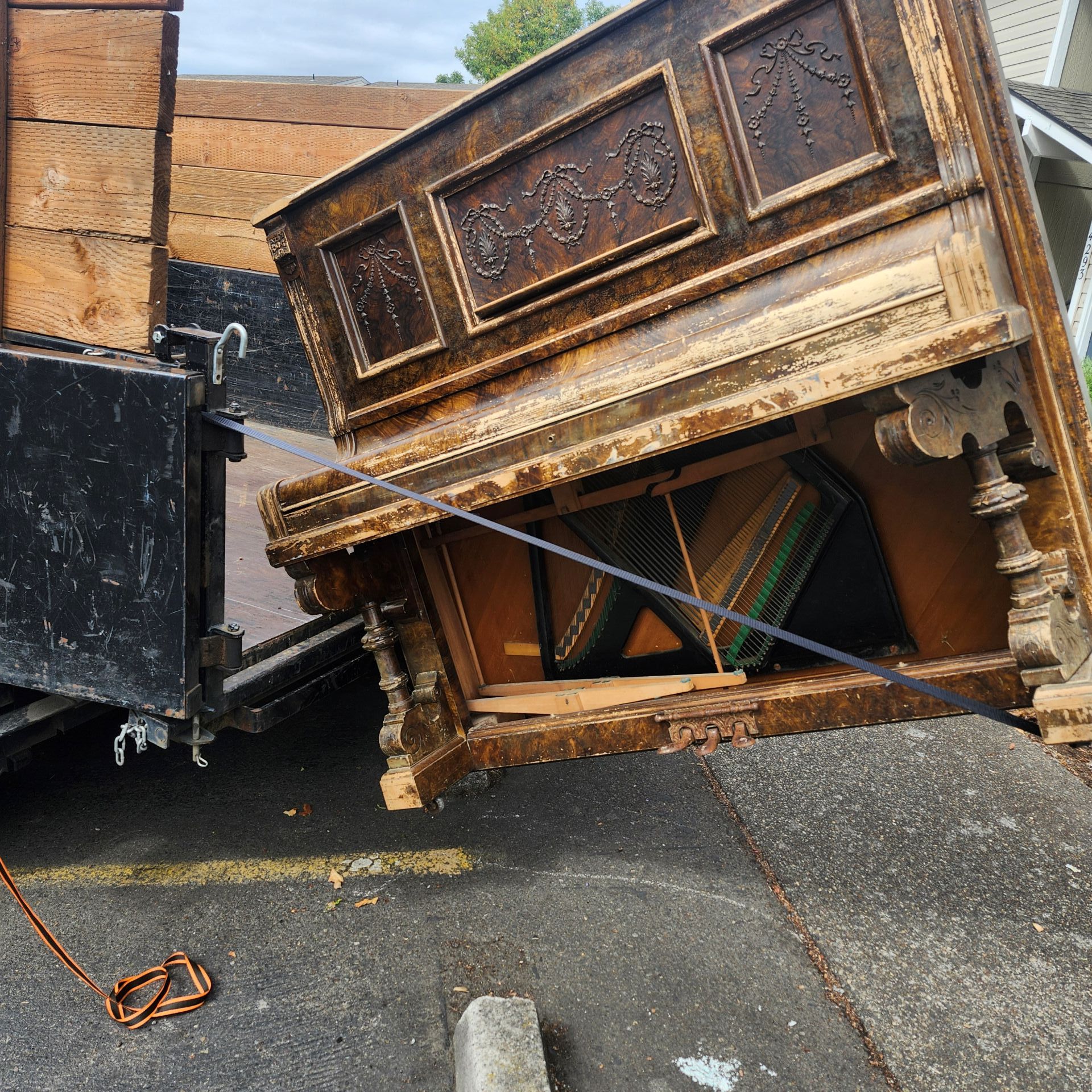 An antique piano being moved, leaning onto a truck bed. Outdoors on asphalt, weathered wood.