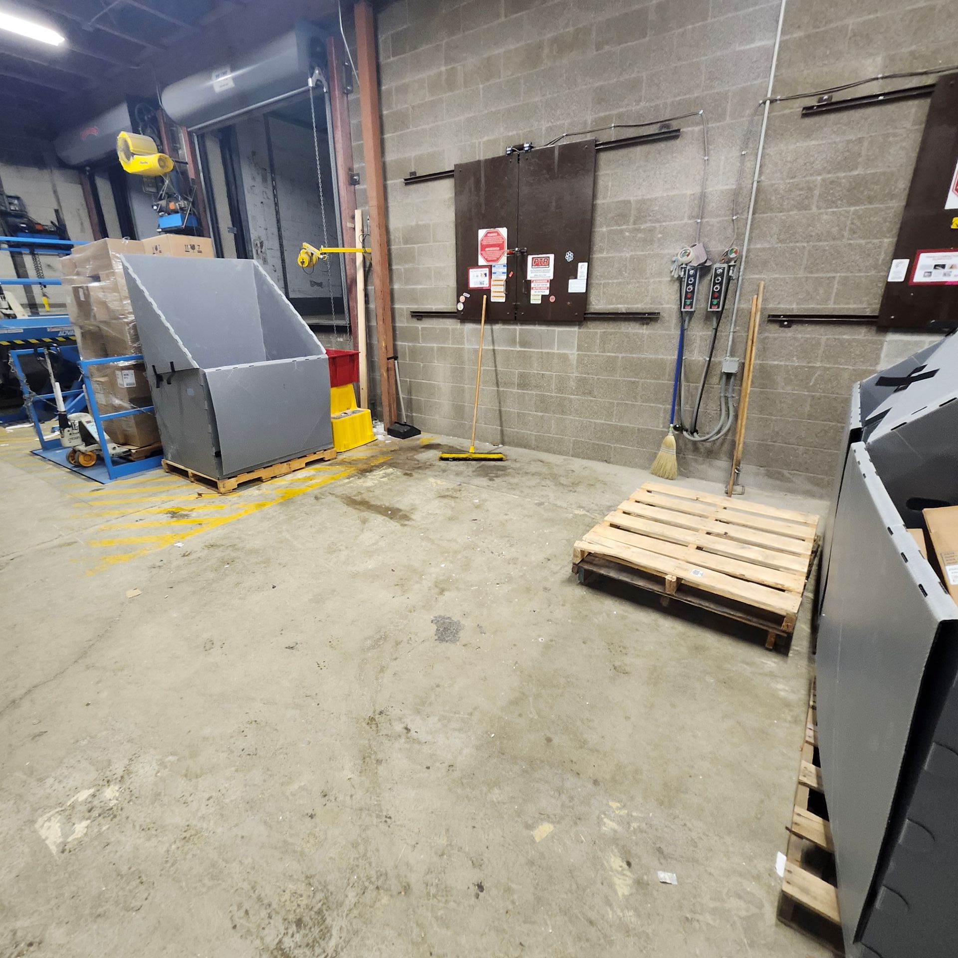 Warehouse interior with a large gray bin, pallet, tools, and signs on a brick wall.