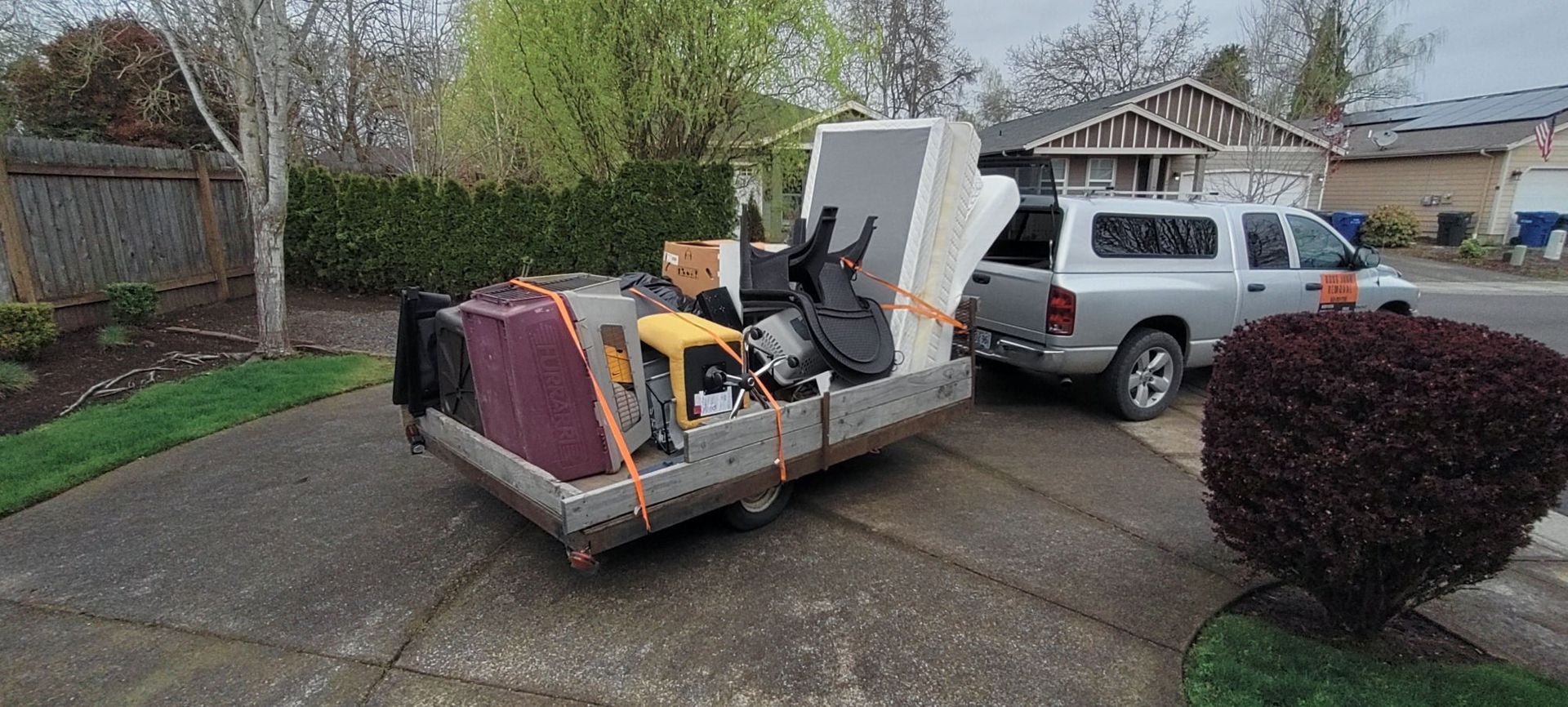 A pickup truck towing a trailer loaded with furniture on a driveway, in front of a house.