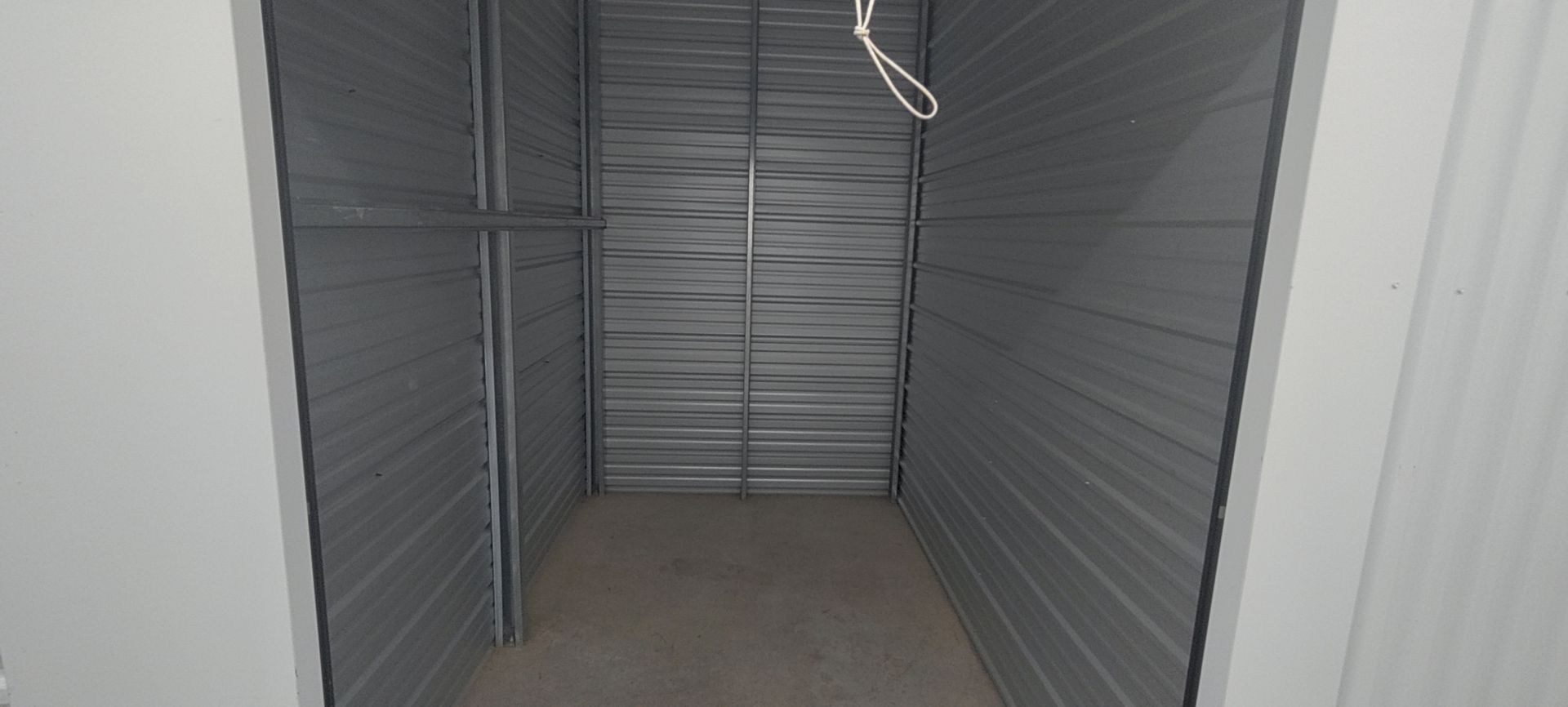 An empty storage unit with gray metal walls and a bare floor. A white cord hangs from the ceiling.