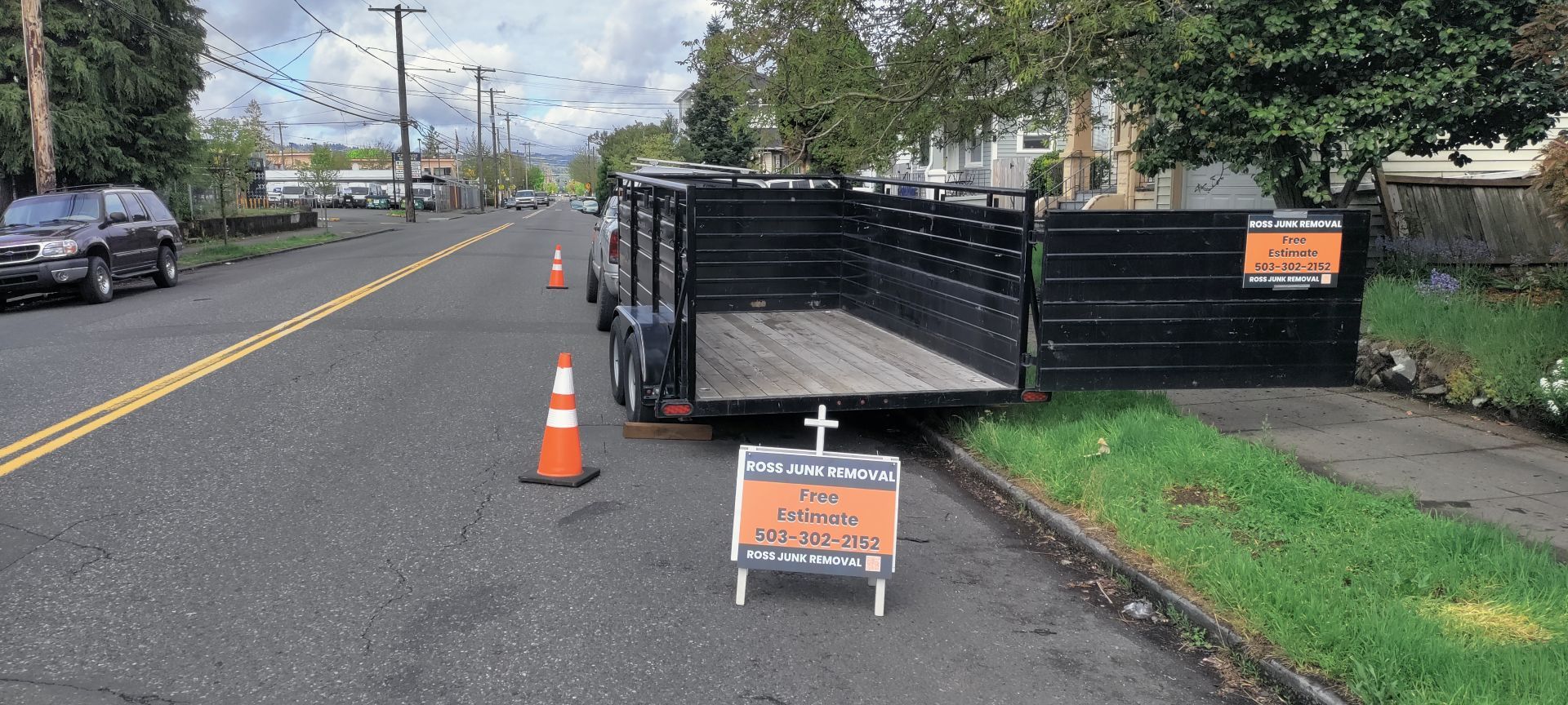 A black trailer parked on a street with orange cones and a sign. A car is parked on the street.