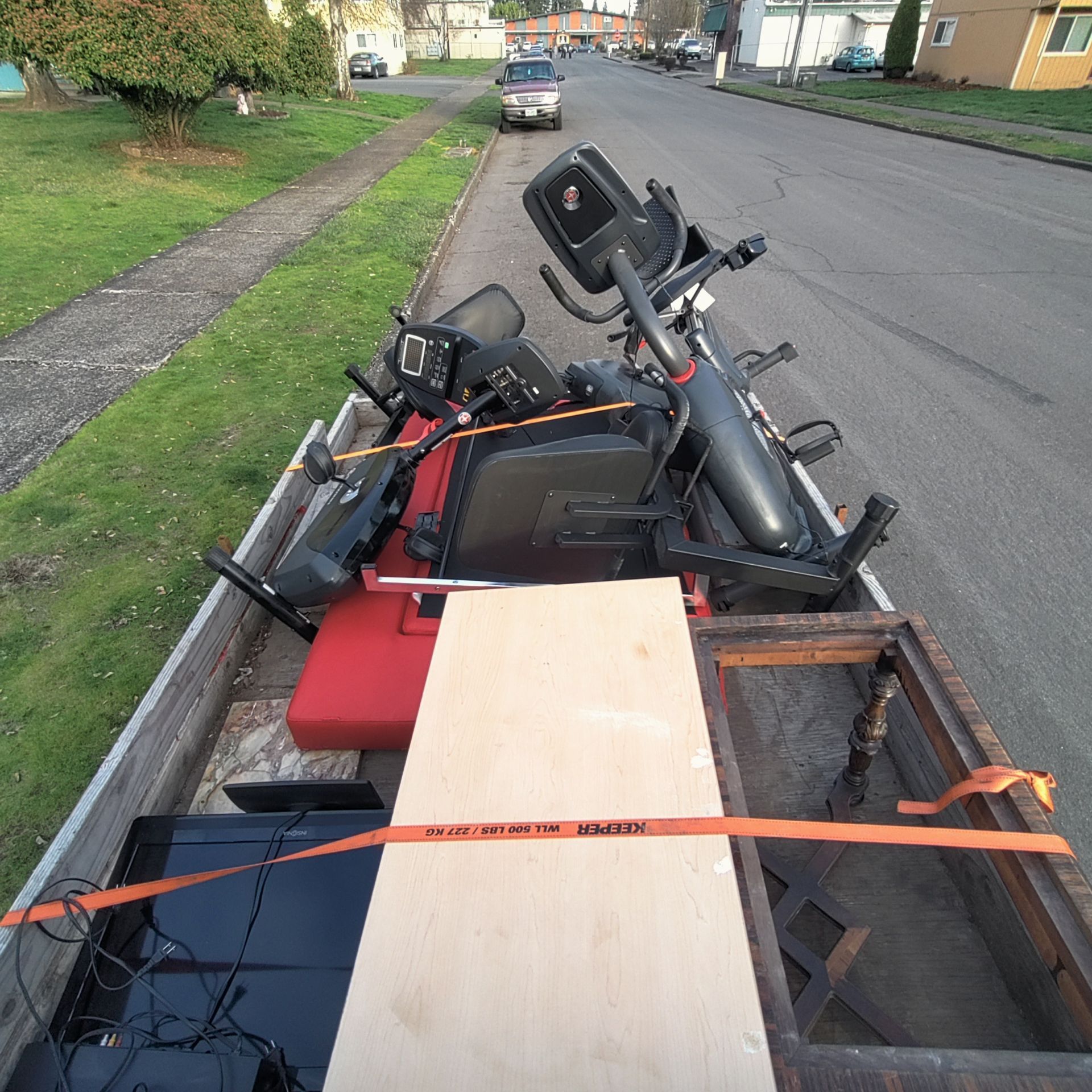 A truck bed filled with assorted discarded items, including exercise equipment and a TV, secured with straps.