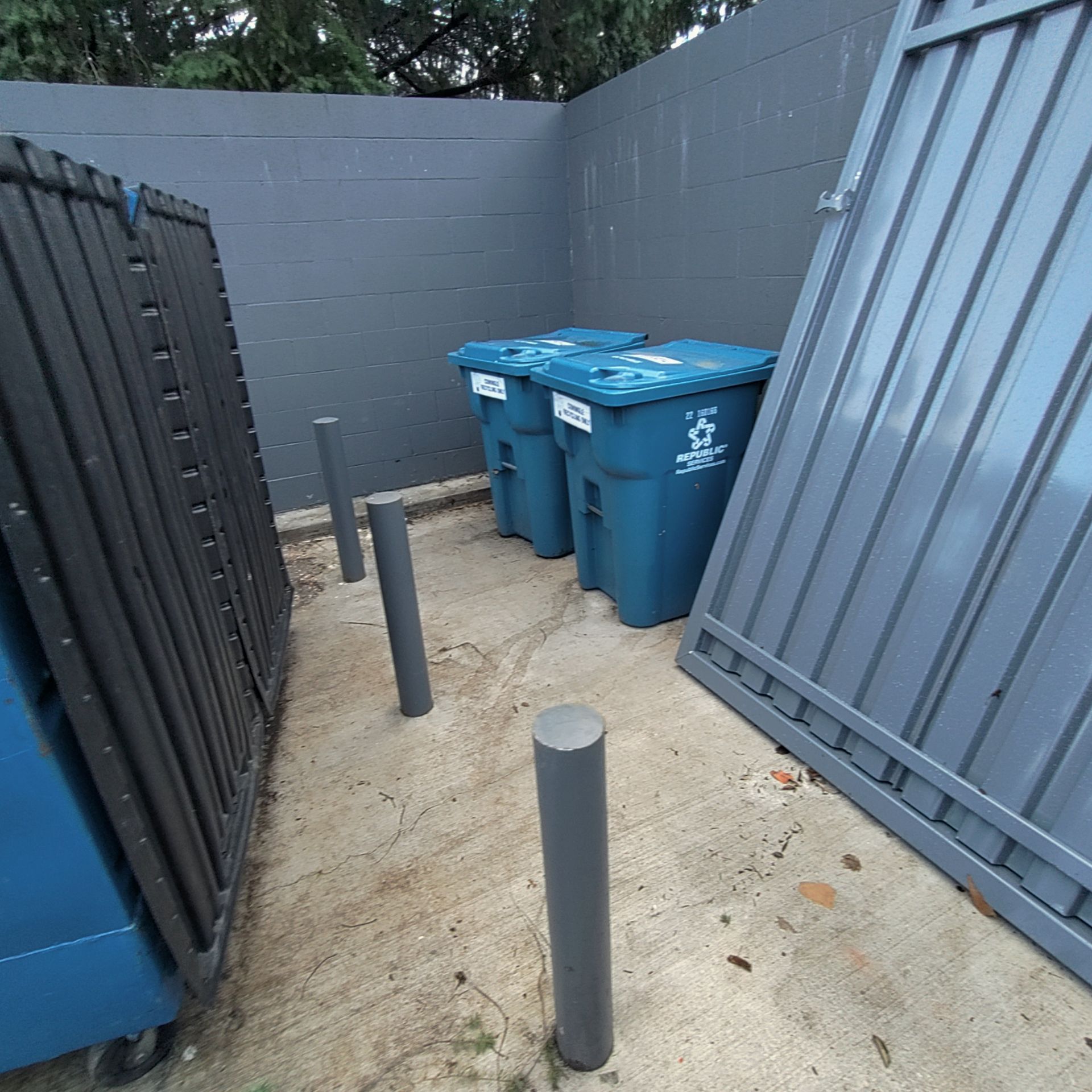 Trash bins in a gray-walled enclosure with a metal gate and concrete bollards.