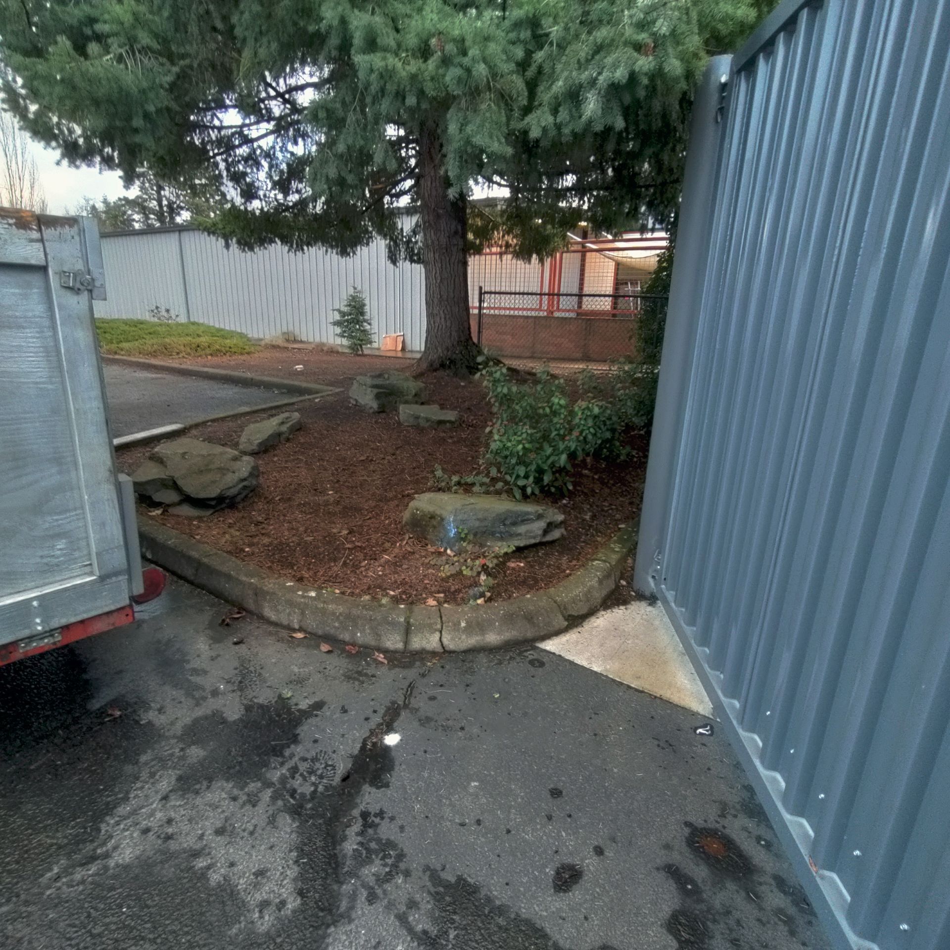 A tree and landscaped area between a metal wall and a parked truck.