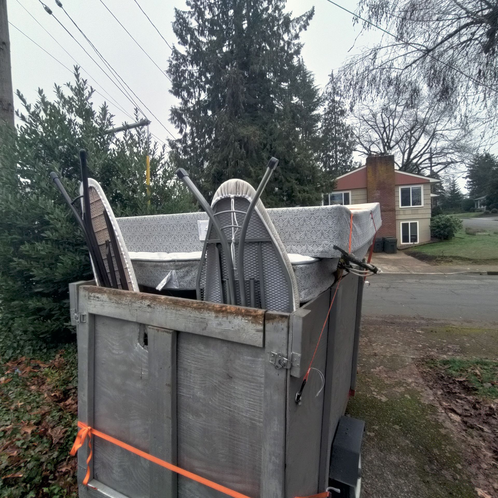 A trailer loaded with various discarded items, including a mattress and chairs, parked on a residential street.