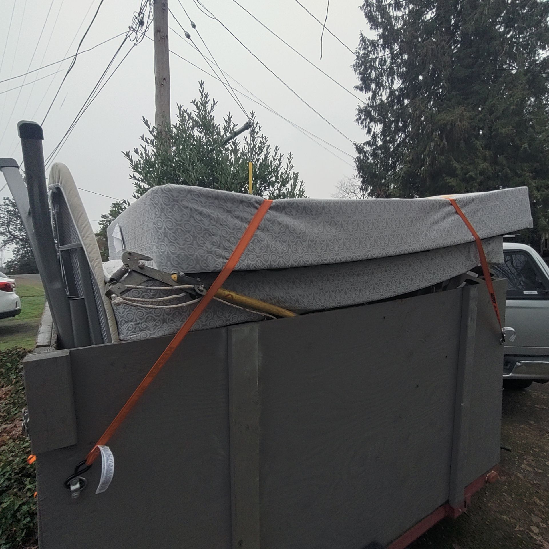 Gray dumpster filled with furniture and metal, secured with orange straps, parked outdoors.