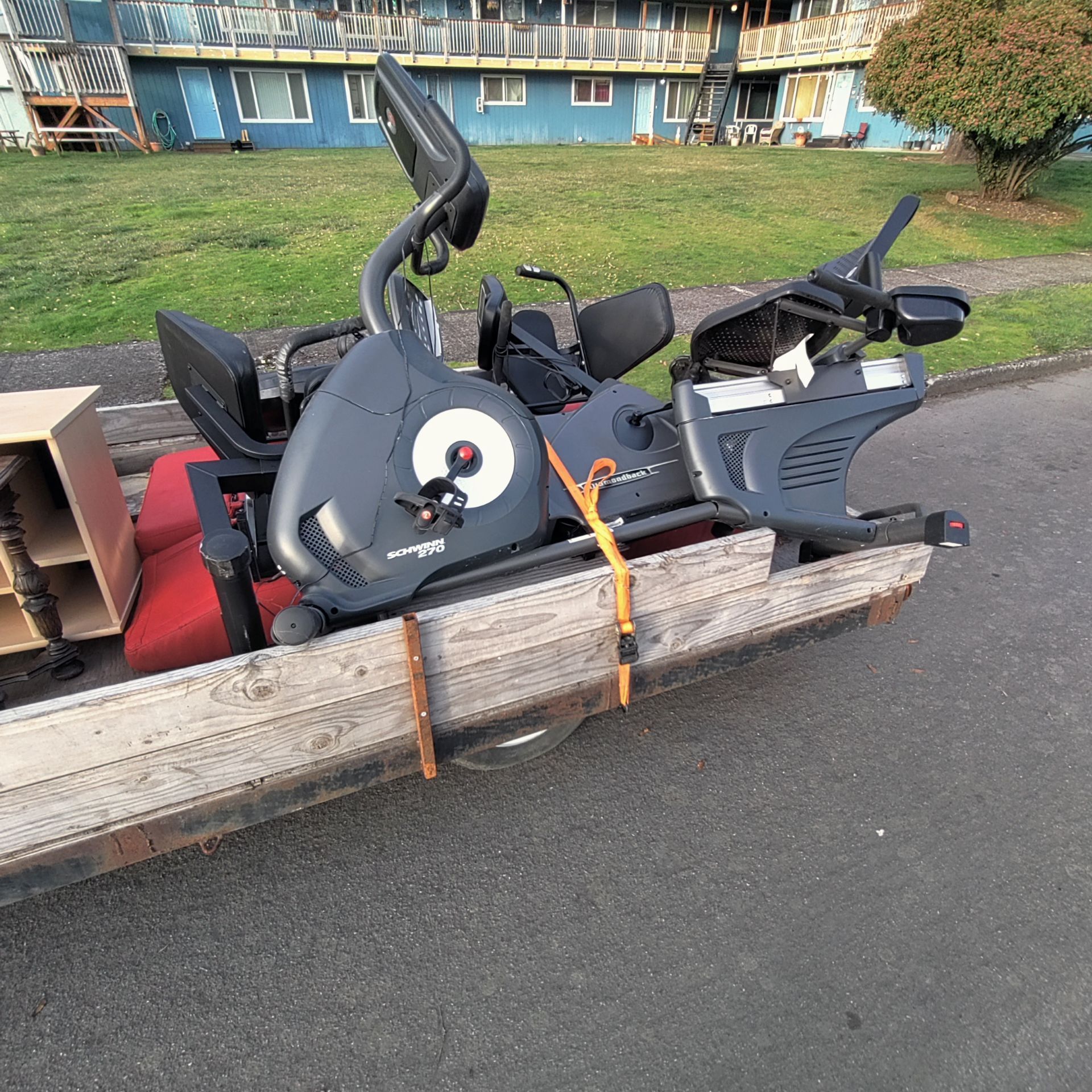 Stationary bike on a wooden frame, secured with straps; outdoors near a blue building on grass.