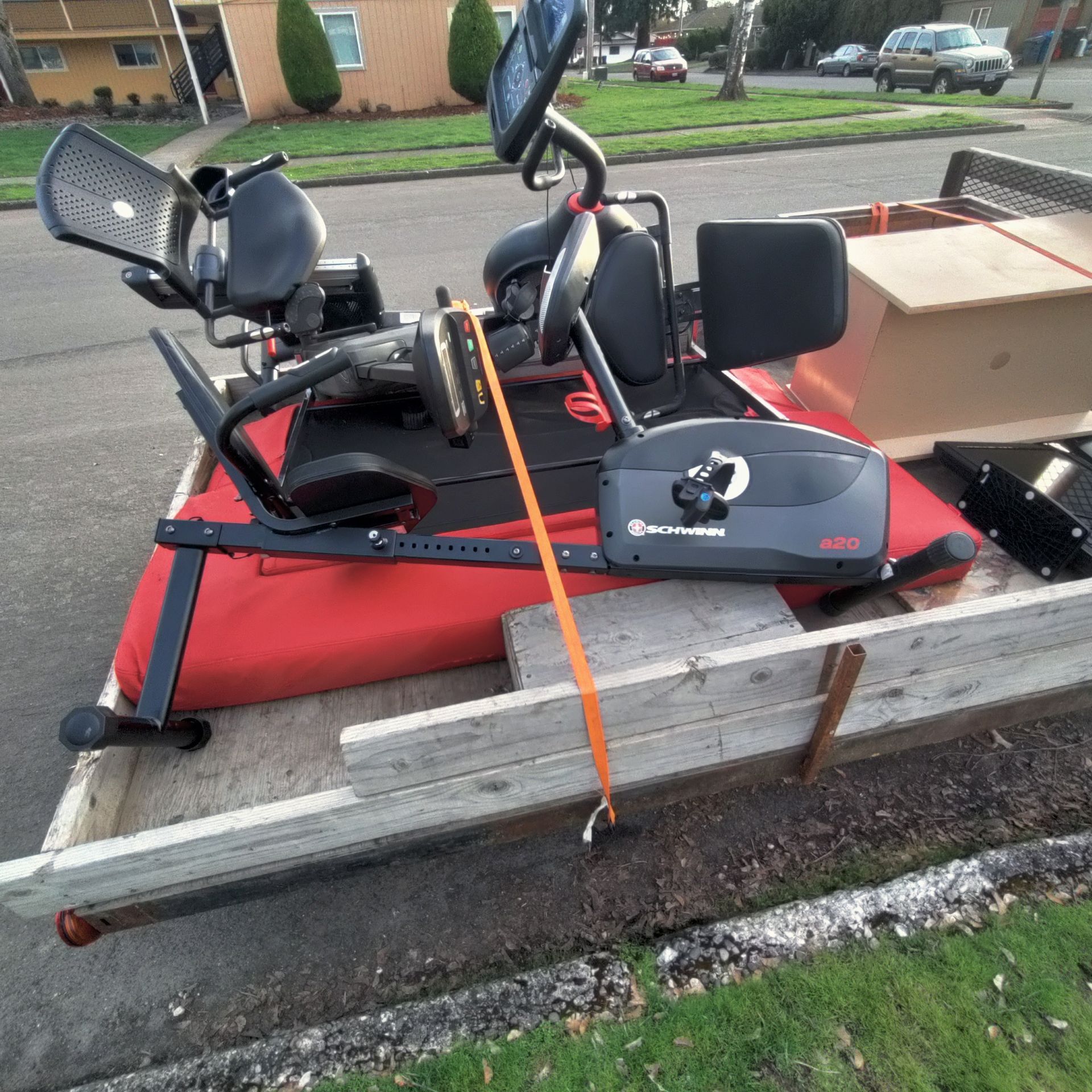 Recumbent exercise bike strapped to a trailer bed, outdoors near a curb and street.