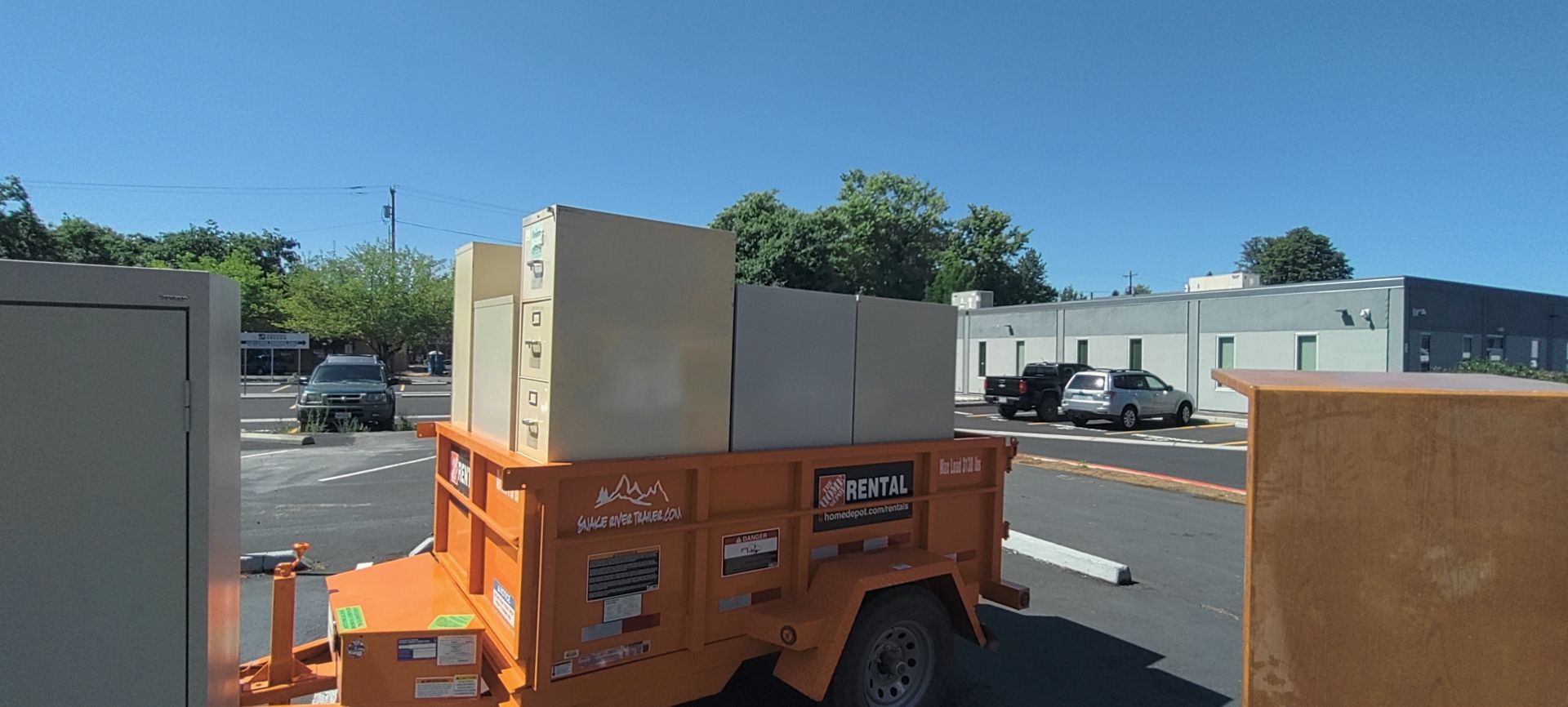 An orange trailer loaded with large boxes parked on a sunny day. Cars and buildings are in the background.