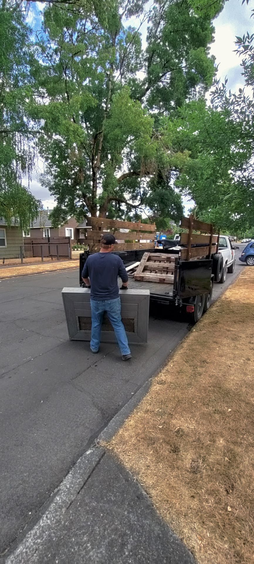 A person loading a large, gray appliance onto a trailer parked on a street lined with trees and houses.