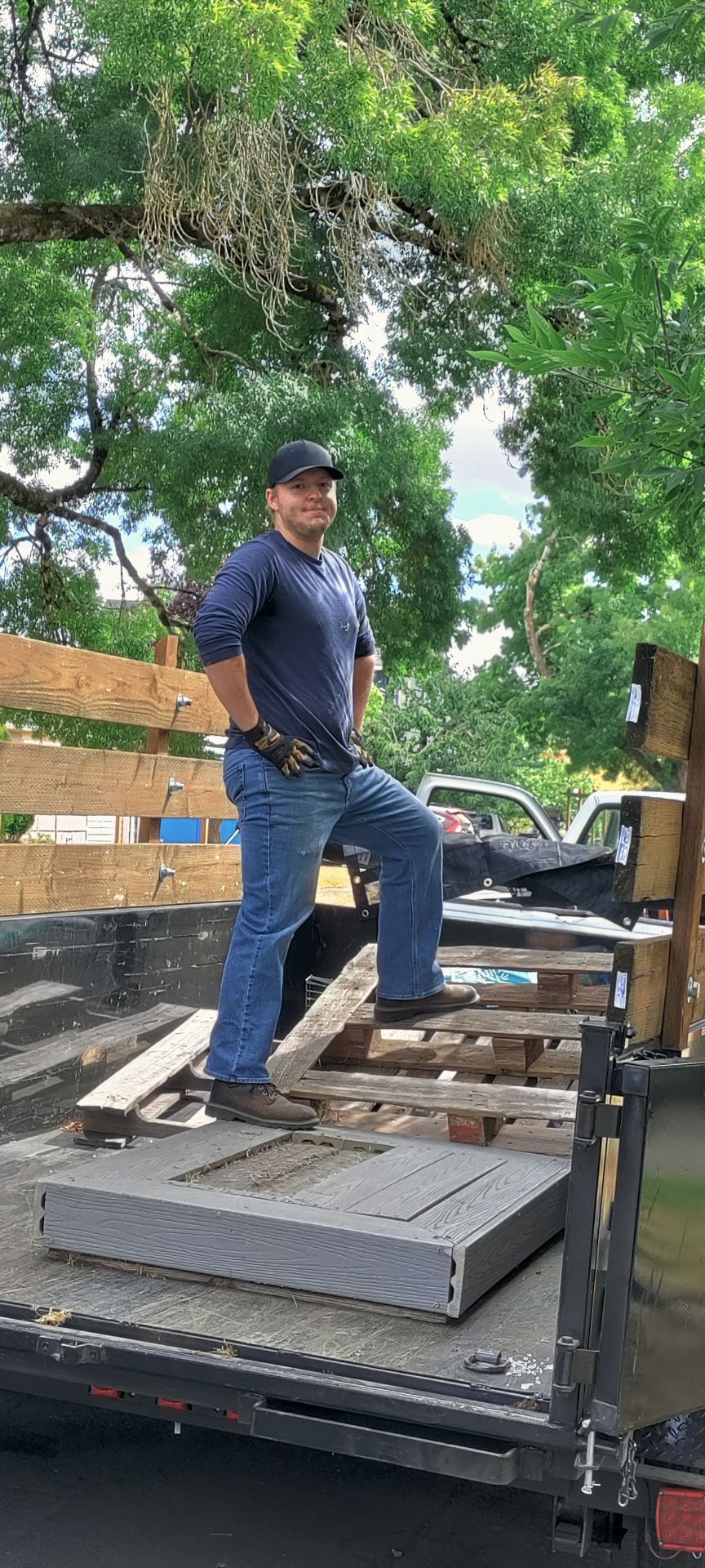 Man standing on a trailer loaded with materials. He wears a hat, jeans, and work gloves. Outdoors.