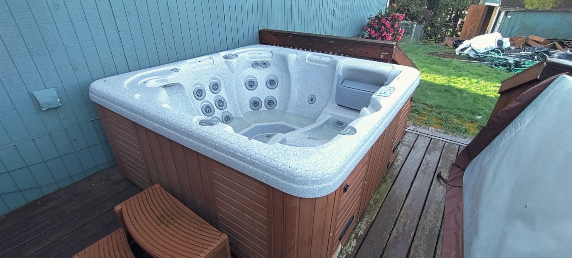 A hot tub sits on a wooden deck next to a grassy area. The hot tub is white with brown siding.