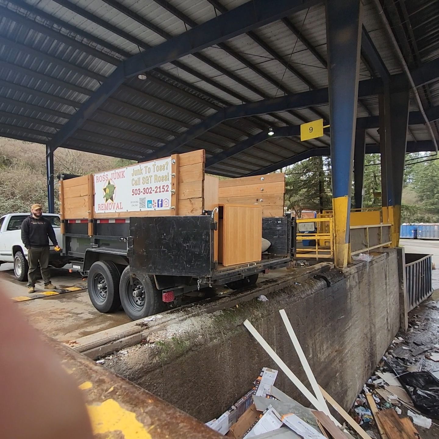Trailer unloading at a waste disposal facility. A man stands beside the trailer, which holds wooden items.