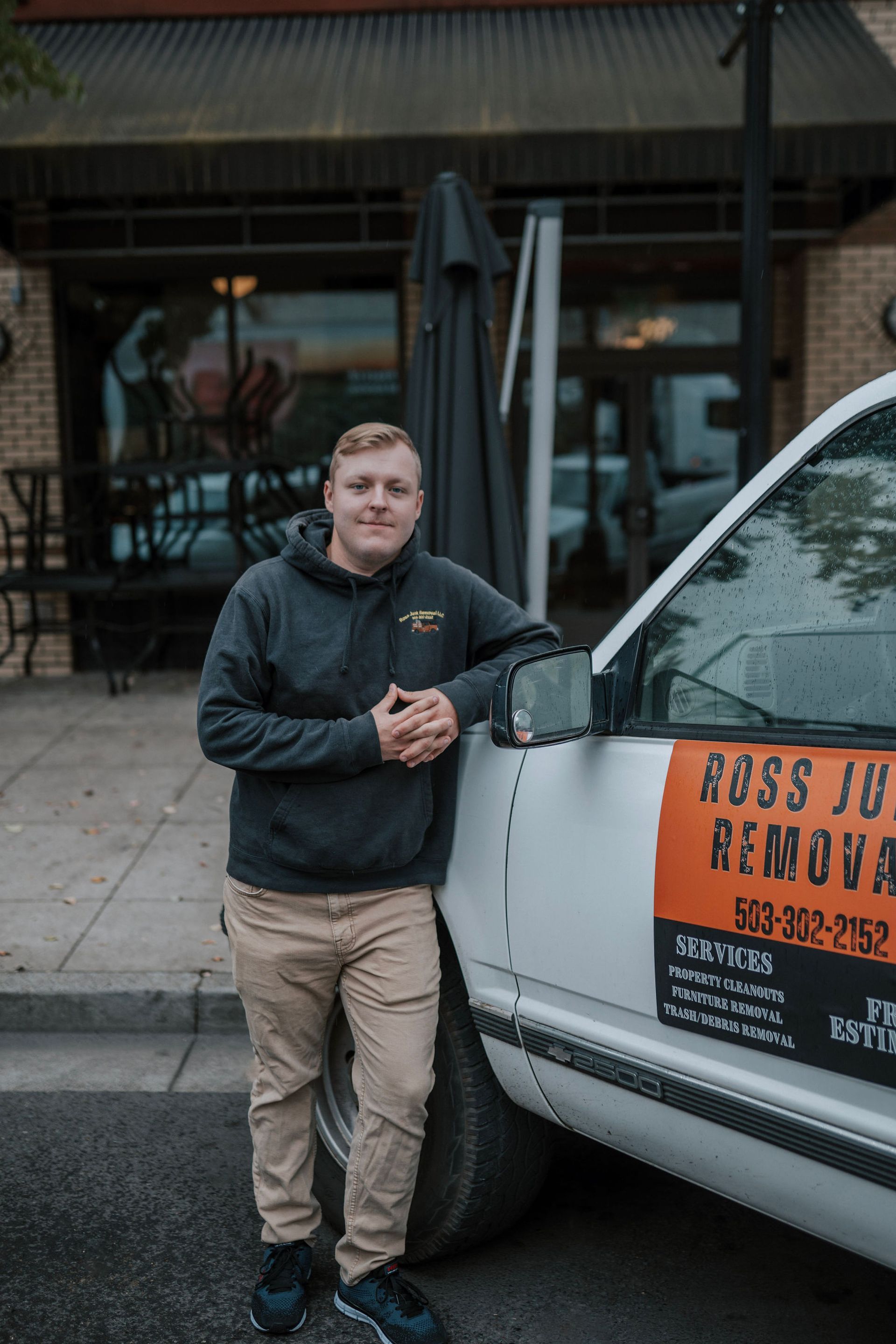 Man in a hoodie and khakis leans on a moving truck with business logo.