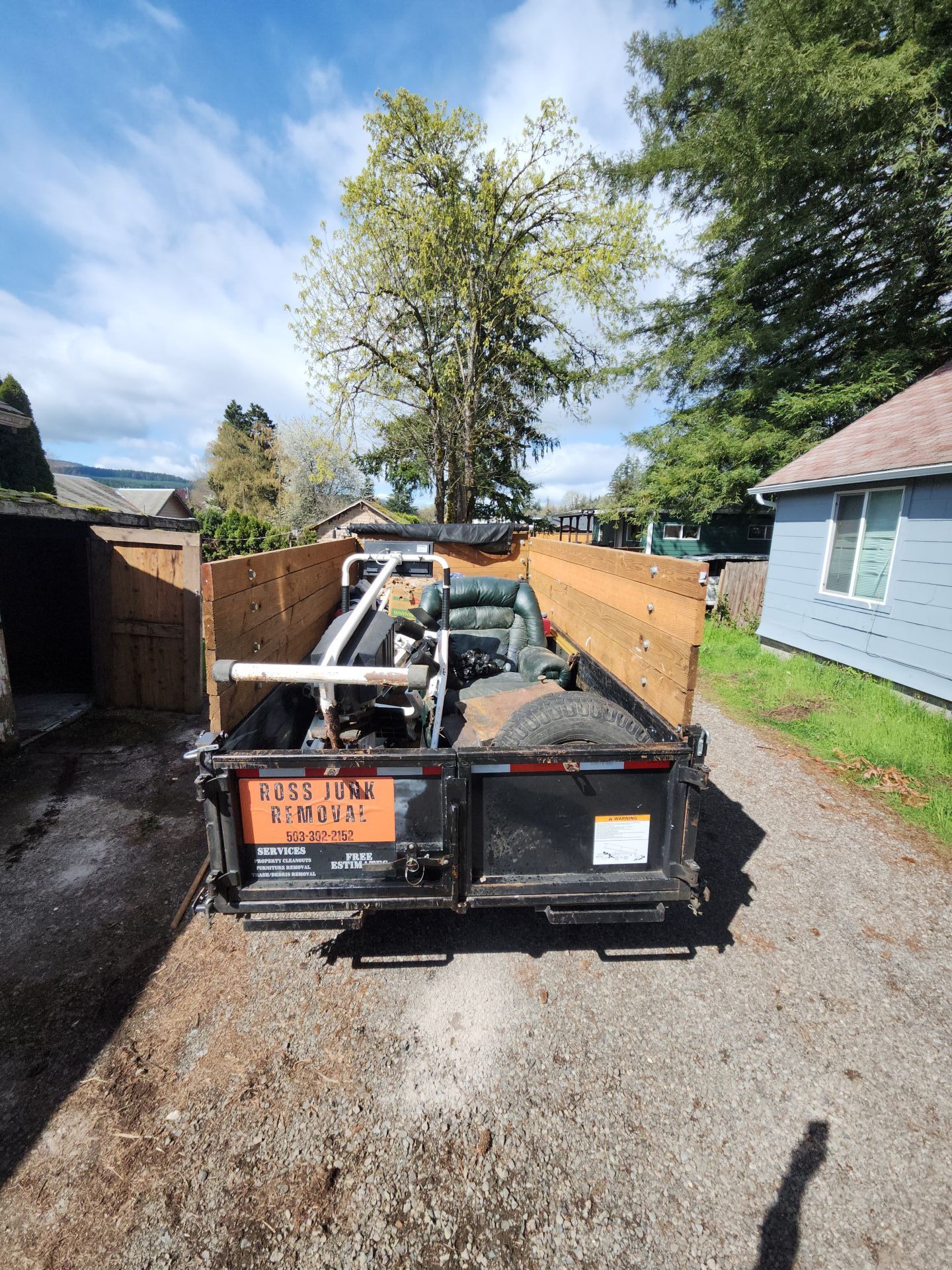 Black trailer filled with scrap metal and debris on a gravel driveway. Wooden fences and a blue house flank it.