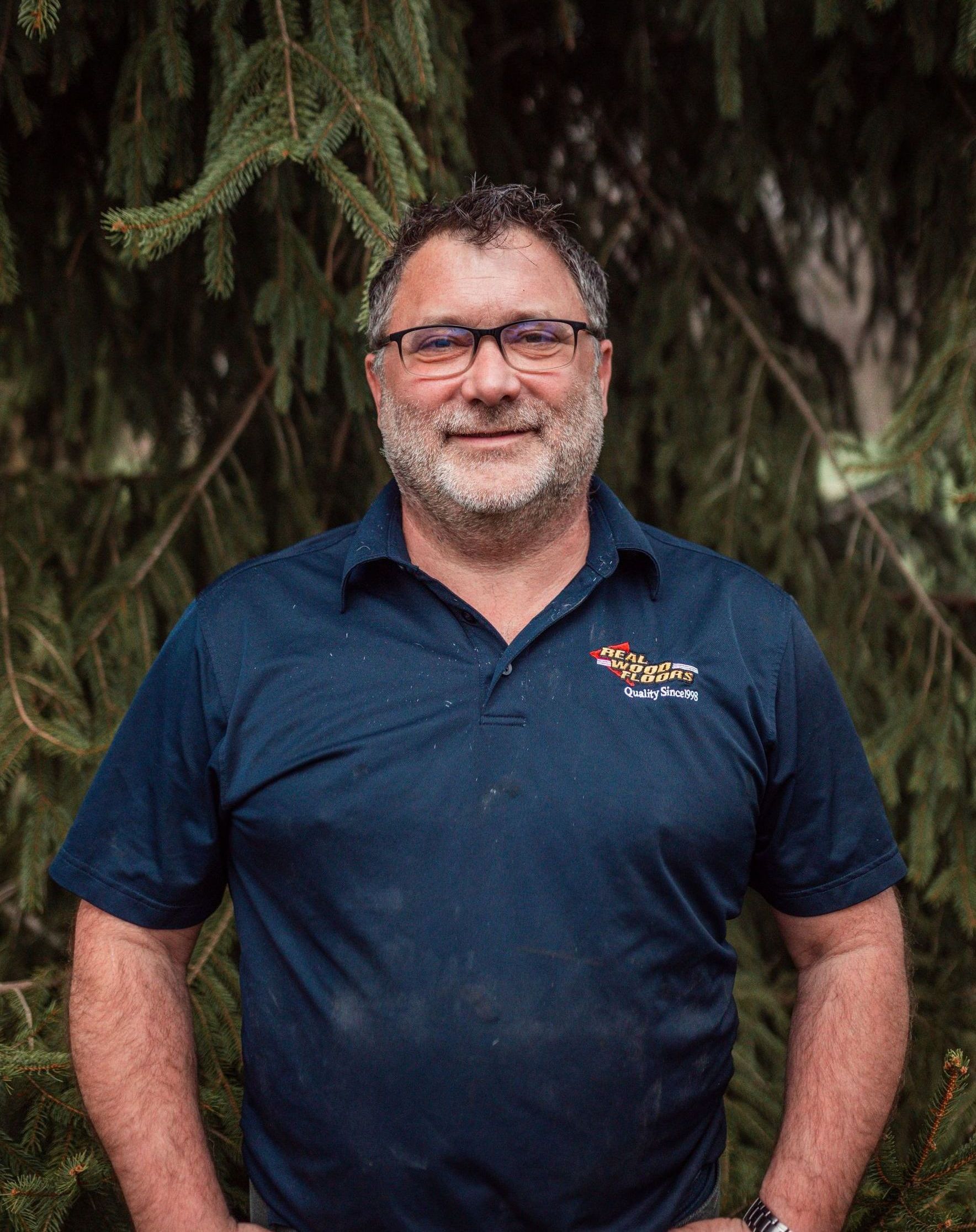 A man with a beard and glasses is standing in front of a tree.