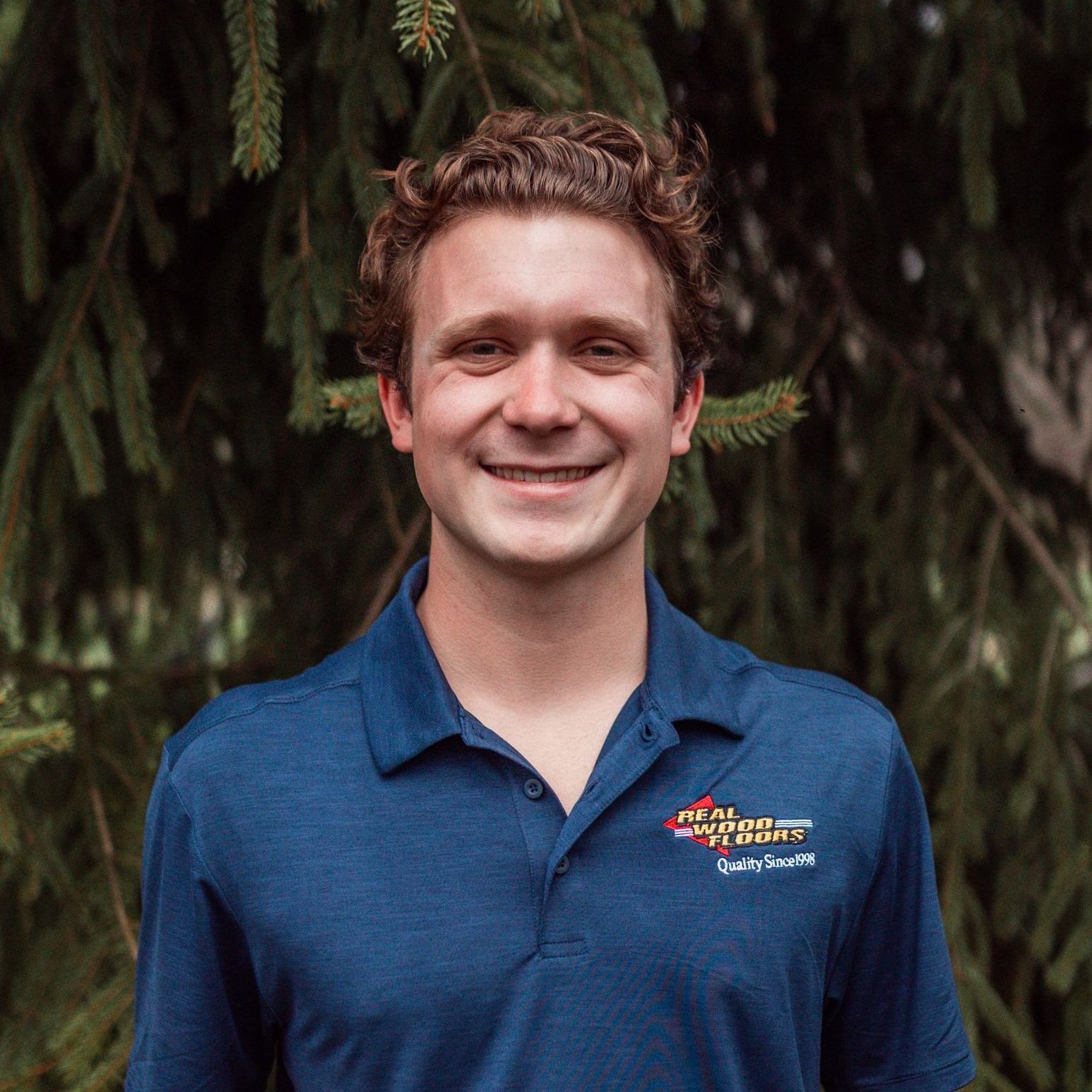 A young man in a blue shirt is smiling in front of a tree.