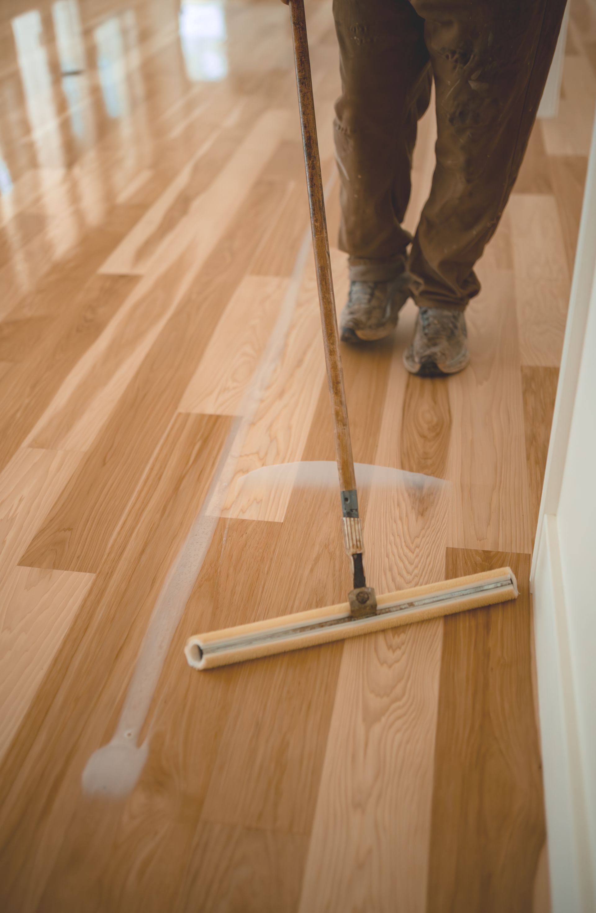 A man is applying finish to a wooden floor with a broom in an empty room.