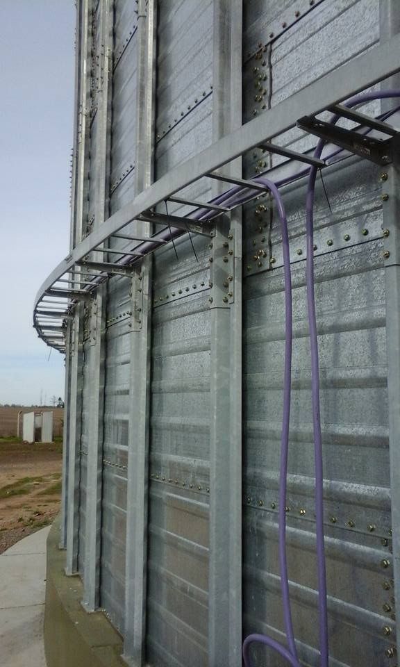 Un silo de grano de metal gris con una escalera curva y cables eléctricos de color púrpura que corren a lo largo del costado.
