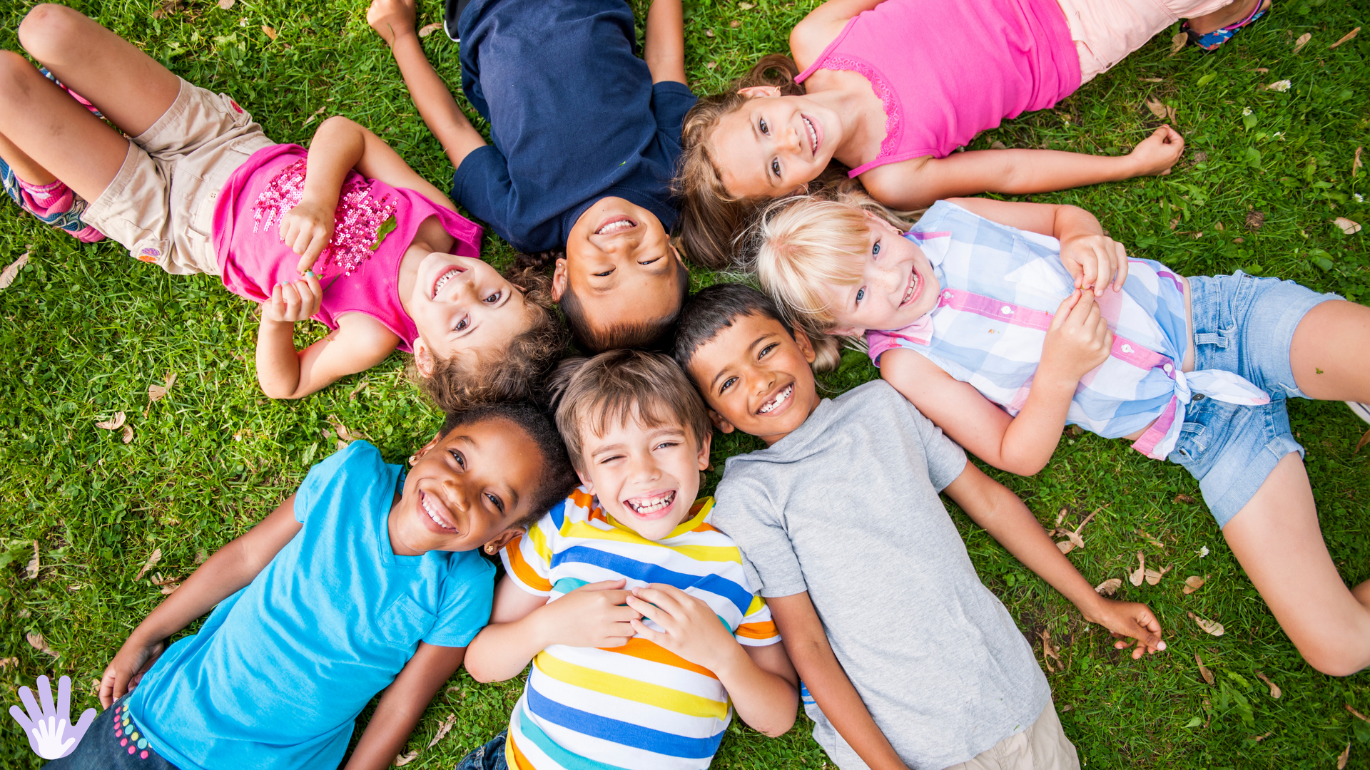 Children lying in a circle on grass, smiling at the camera.
