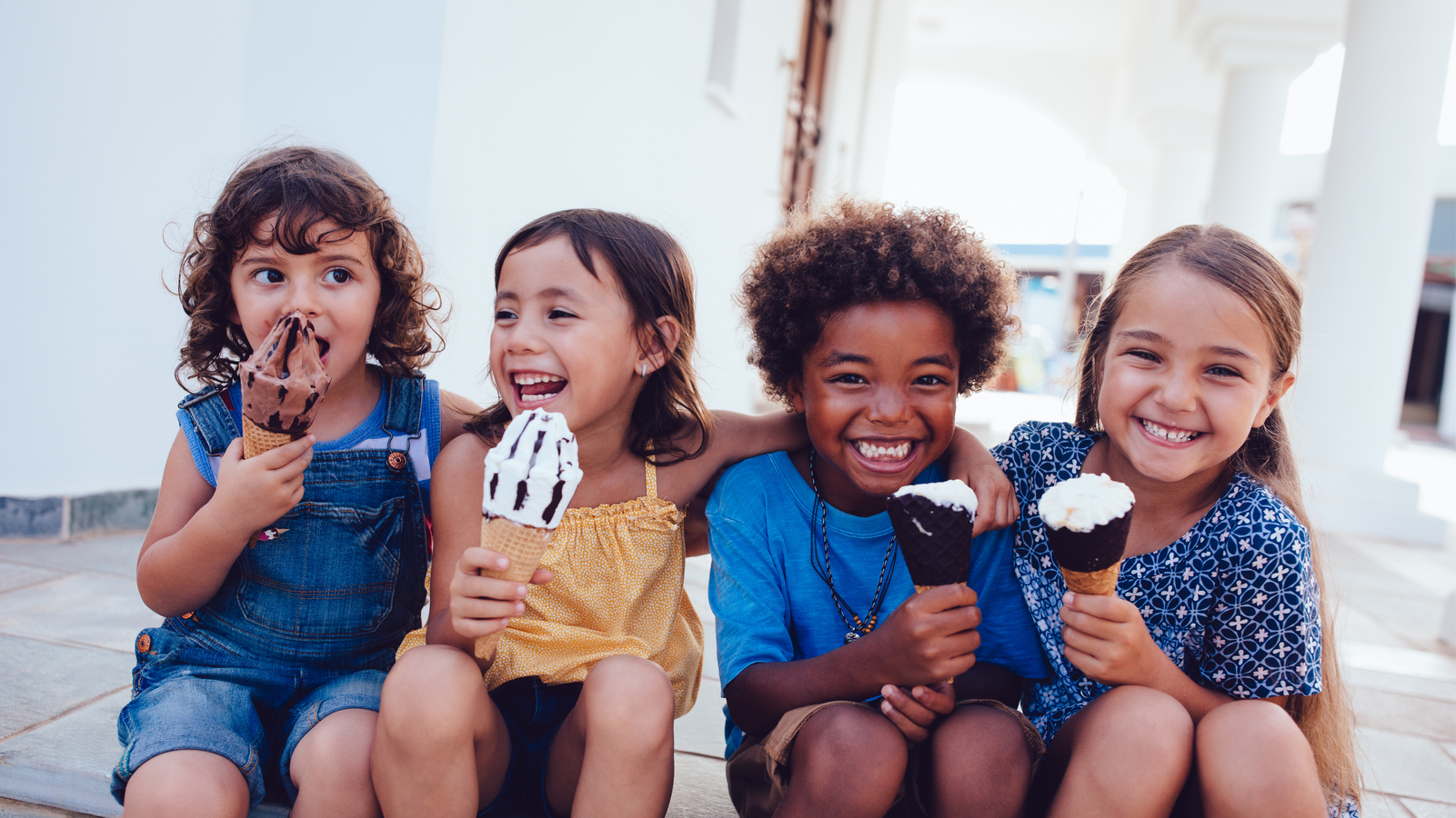 Four children sitting together, smiling while eating ice cream cones in an outdoor setting.