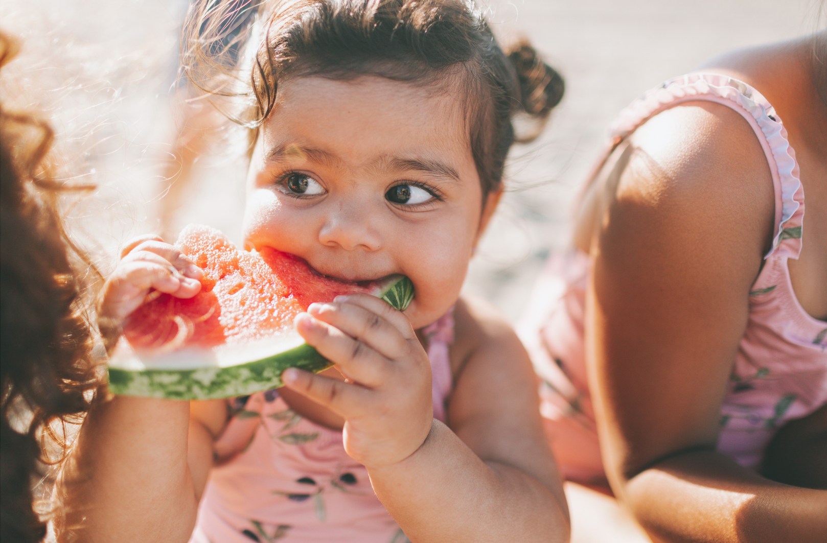 A young child with pigtails holding and eating a slice of watermelon while wearing a pink patterned swimsuit.