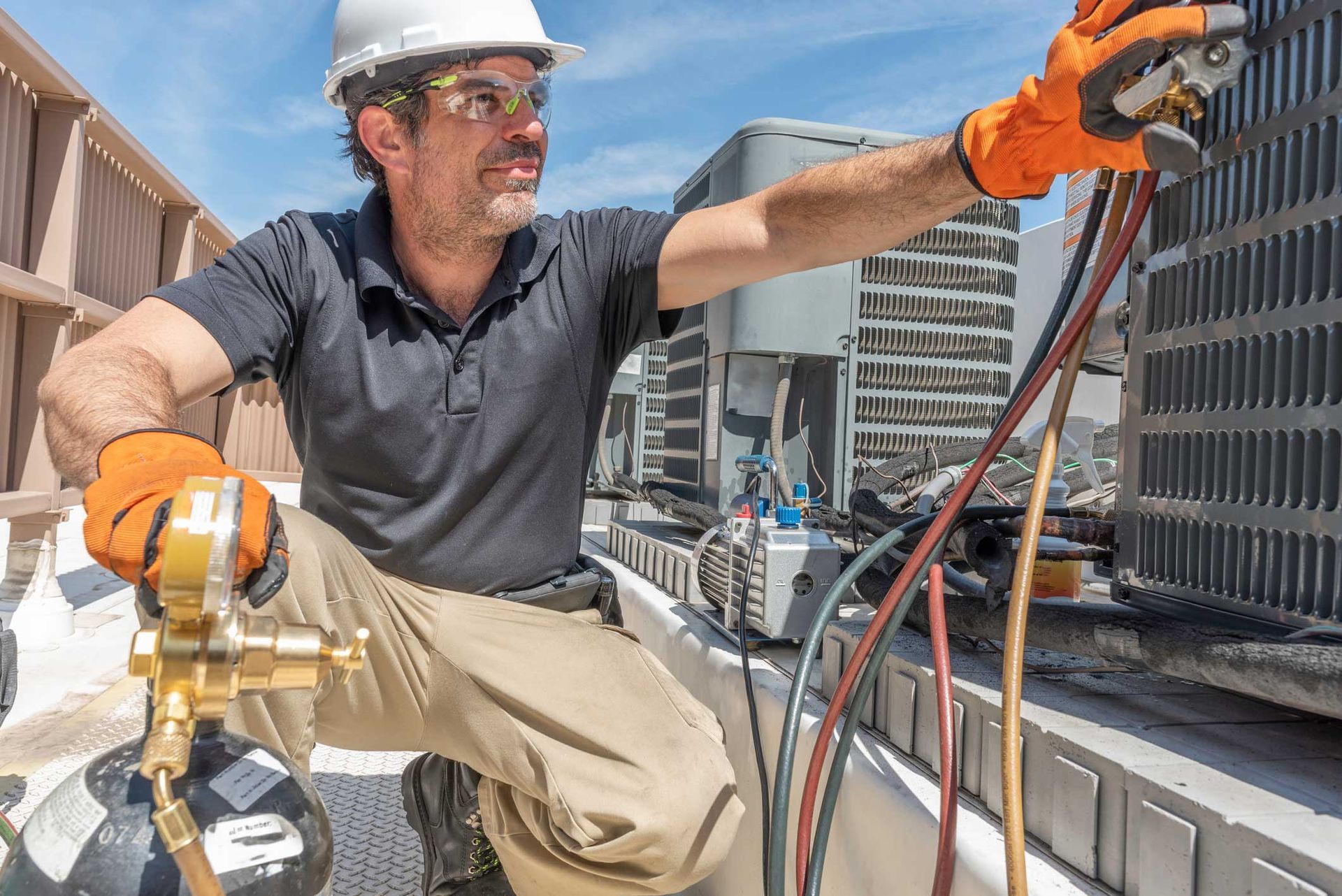 HVAC technician works on rooftop unit, wearing a hard hat and safety glasses, with hoses and gauges.