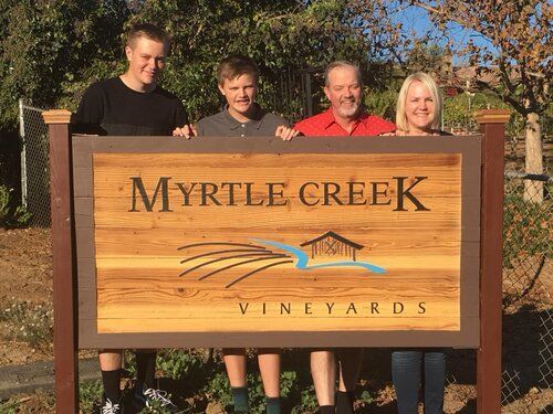 A family is standing next to a wooden sign that says myrtle creek vineyards.