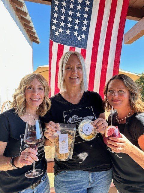 Three women are standing in front of an american flag holding wine glasses and snacks.