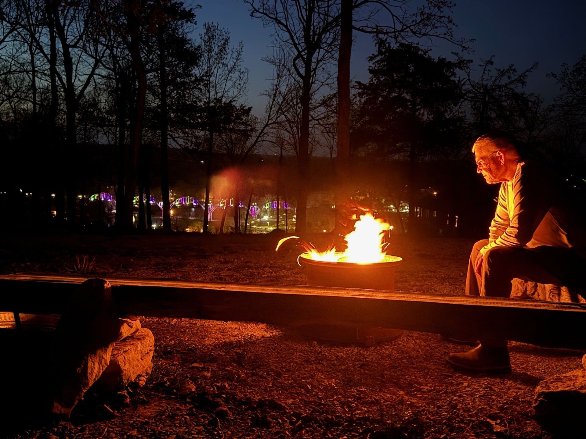 Person sitting by a bonfire at night. Flames illuminate their face and surrounding trees. Dark sky.