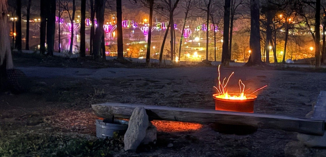Night scene: a campfire burns in a fire pit, with lights of a town visible in the distance through trees.