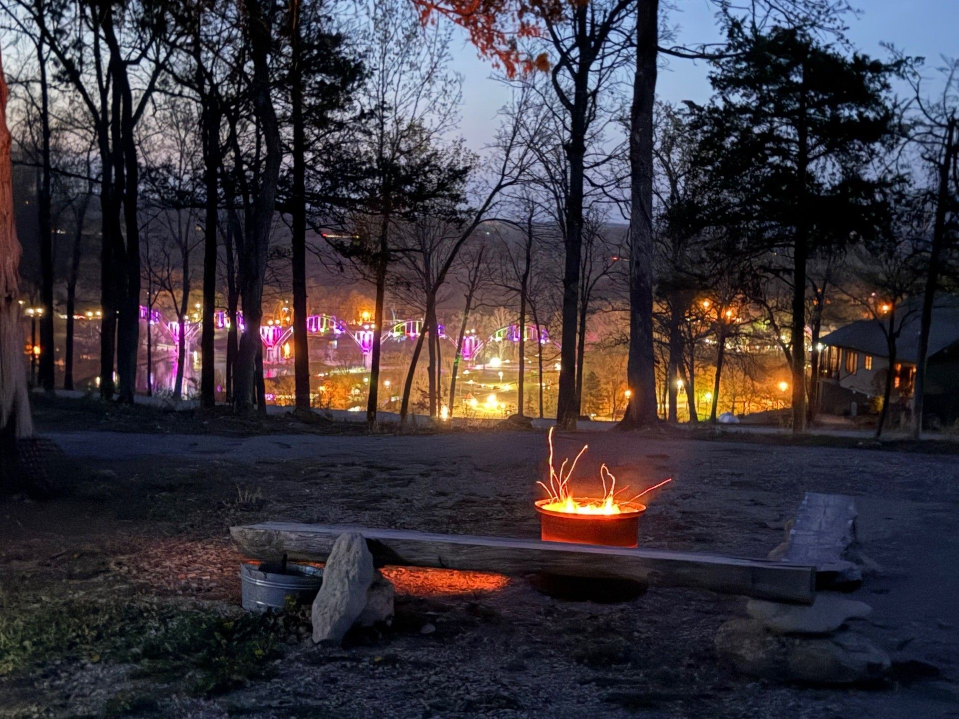 A campfire burns in front of a bench, with city lights in the distance through trees at dusk.