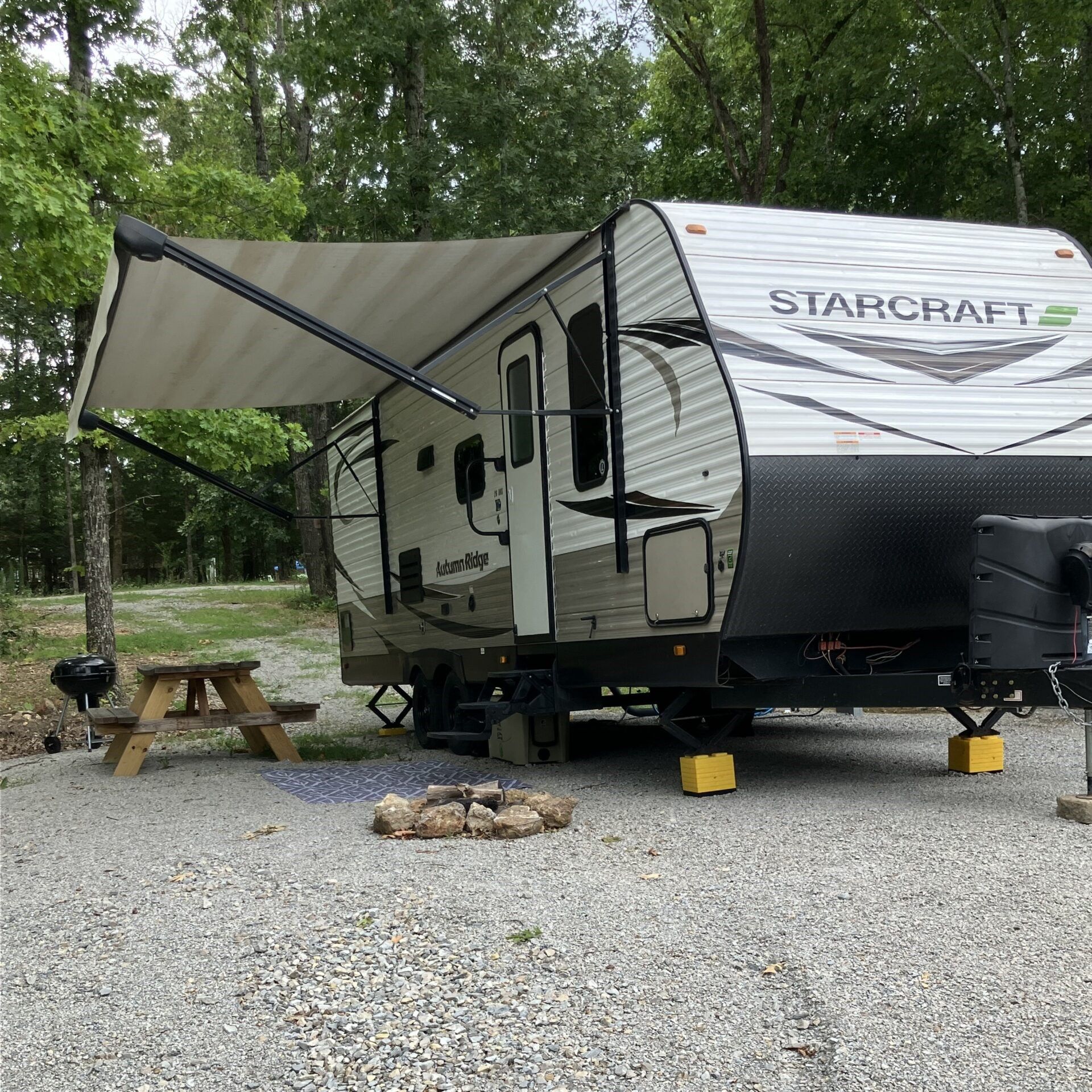 A Starcraft RV with awning extended, parked in a gravel campsite next to a picnic table and fire pit.