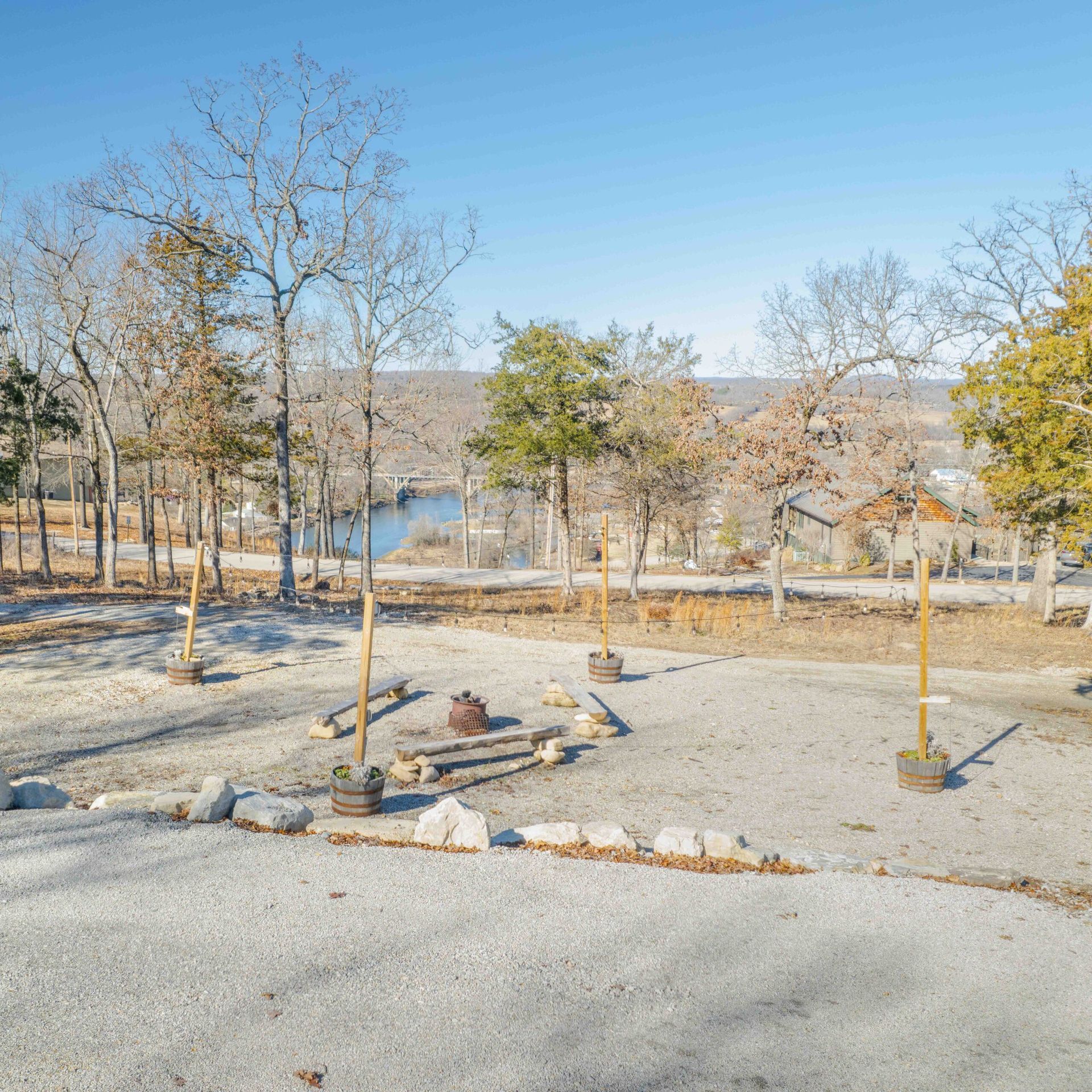 A gravel clearing with a fire pit, surrounded by trees under a bright blue sky, overlooking a lake.